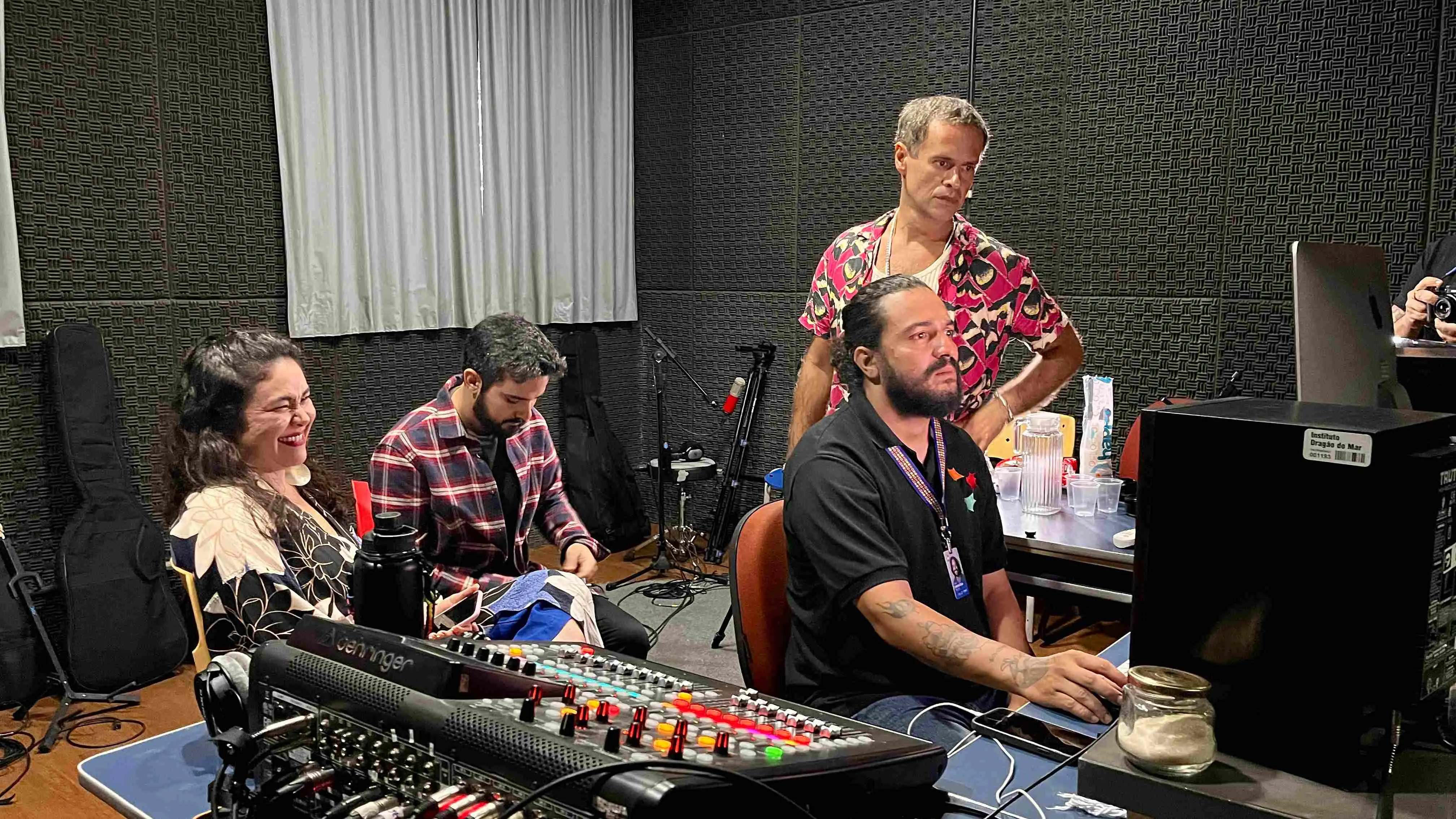 Em um ambiente de estúdio de gravação profissional, com paredes forradas em espuma acústica preta, quatro pessoas interagem em torno de uma grande mesa de som Behringer que domina o primeiro plano. Sentada à esquerda, uma mulher de blusa floral estampada sorri, olhando para fora da cena, enquanto um homem ao centro, de camisa xadrez em tons de vinho, está inclinado e concentrado em algo no colo. À direita, um homem de barba, cabelos longos e camisa polo preta está sentado e focado na mesa de som e no computador, manipulando os equipamentos. Atrás dele, em pé, está Marcelo Jeneci – vestindo sua camisa de estampa vibrante e colorida –, que observa a cena com as mãos na cintura e uma expressão séria, sugerindo um momento de tutoria ou ajustes técnicos do grupo.