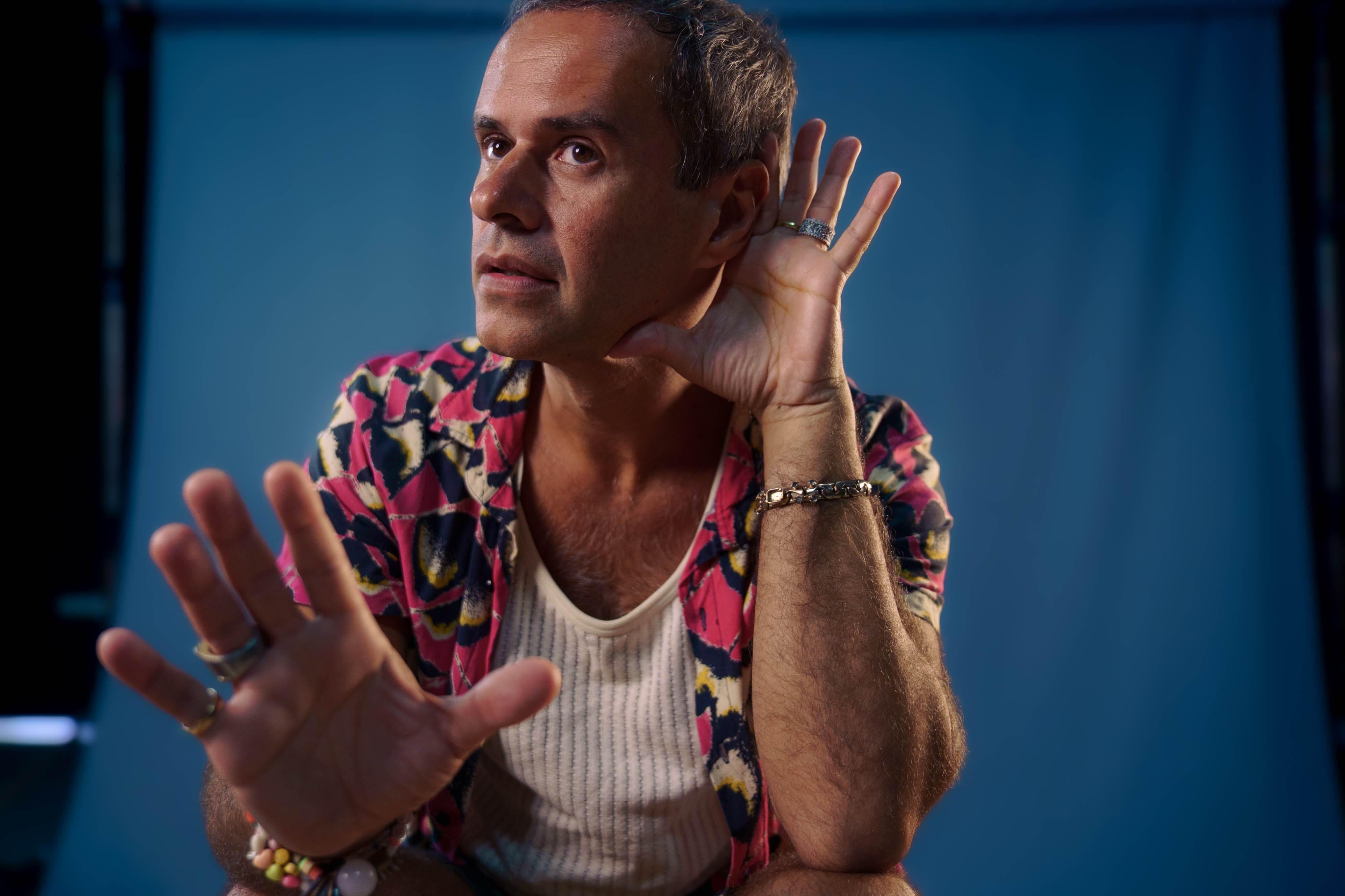 Em um plano médio e com iluminação dramática contra um fundo azul escuro, o músico e cantor Marcelo Jeneci está em foco, exibindo uma expressão de intensa escuta ou atenção, com os olhos fixos para a frente, sugerindo que ele está captando algo no ambiente ou pedindo silêncio. Ele veste uma camisa de botão de manga curta com estampa vibrante em tons de rosa, vermelho e preto sobre uma camiseta regata branca. Seus braços e mãos estão em destaque: a mão direita está levantada ao lado da cabeça, com a palma ligeiramente virada para o ouvido em um gesto de 