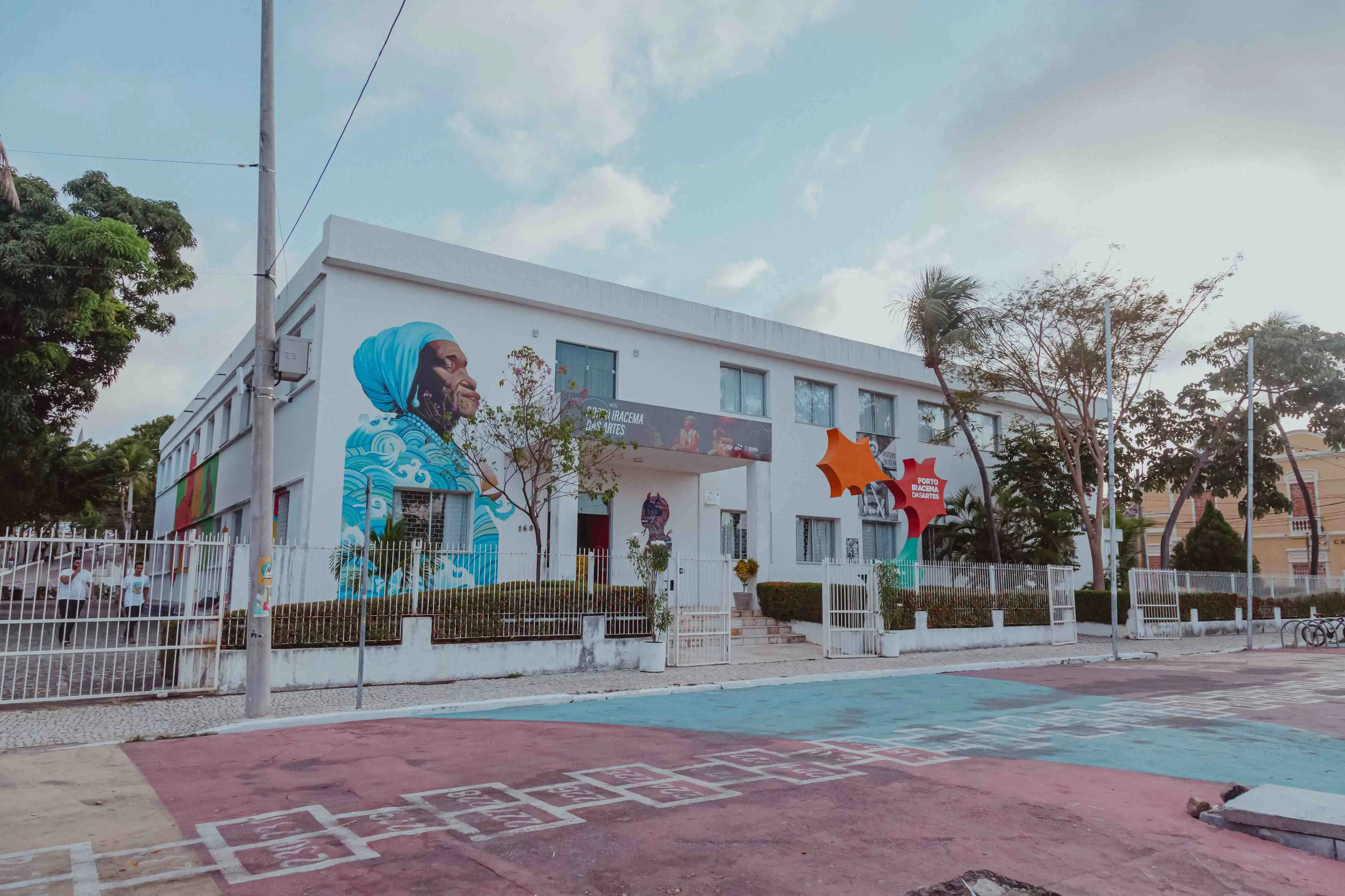 A imagem é uma foto de um edifício de dois andares, de cor branca, com uma fachada que apresenta vibrantes murais de arte. O edifício parece ser uma instituição cultural ou de artes, talvez identificada pelo texto na fachada. Um grande e colorido mural domina o lado esquerdo, retratando o perfil de uma pessoa mais velha, possivelmente uma mulher, usando um turbante azul e vestindo roupas com padrões azuis e brancos. À direita da entrada principal, que tem uma pequena placa de identificação vermelha e branca, há outro mural colorido e abstrato, com formas geométricas em tons de laranja, vermelho e verde, incluindo um elemento em forma de estrela ou flor de quatro pontas. Em frente ao edifício, há uma cerca branca de ferro, e além dela, o chão da calçada é pintado em faixas largas e coloridas de azul, rosa, e tons terrosos, com o desenho de uma amarelinha em primeiro plano, sugerindo um espaço público convidativo. Há algumas árvores e arbustos verdes que emolduram o edifício na frente e nos lados, e o céu acima é parcialmente nublado com nuvens brancas e azuis. A cena é bem iluminada, indicando que a foto foi tirada durante o dia.