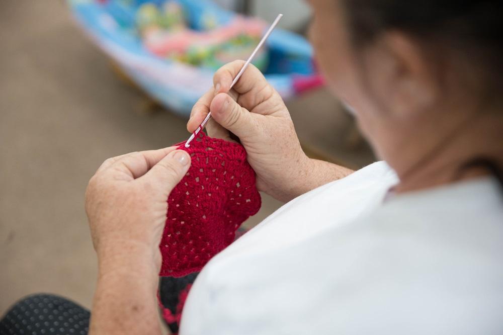Na imagem, close-up sobre o ombro de uma mulher mais velha, com o foco em suas mãos trabalhando em uma peça de crochê vibrante na cor vermelha com um gancho de crochê rosa claro. Ela está vestindo uma camiseta branca e a peça de crochê possui pontos abertos.