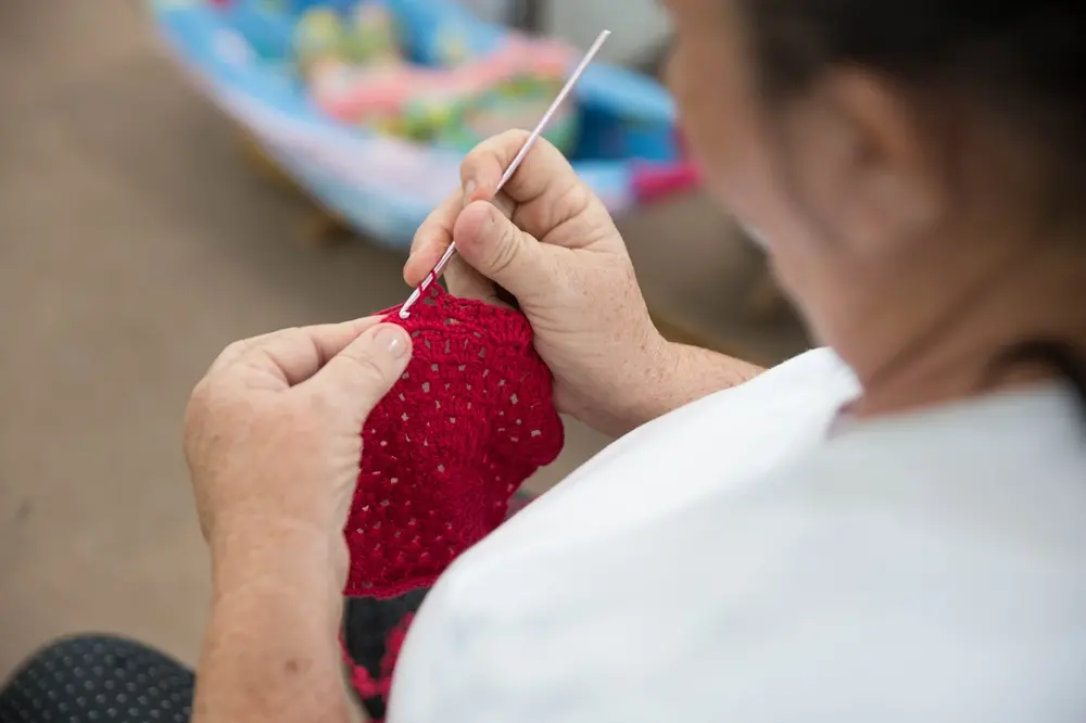 Na imagem, close-up sobre o ombro de uma mulher mais velha, com o foco em suas mãos trabalhando em uma peça de crochê vibrante na cor vermelha com um gancho de crochê rosa claro. Ela está vestindo uma camiseta branca e a peça de crochê possui pontos abertos.