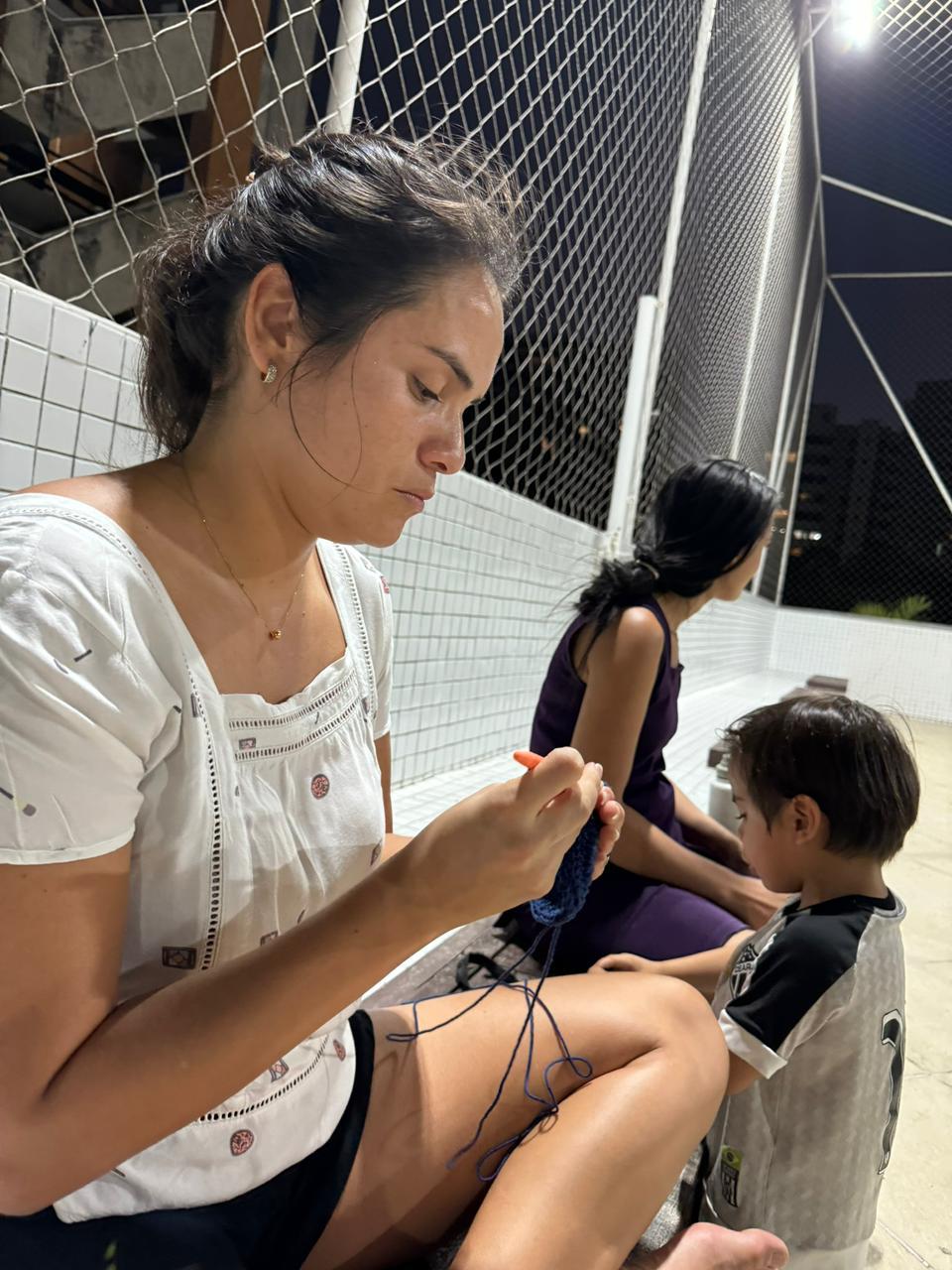 Na imagem, mulher sentada, Cinara Eufrásio, com cabelos castanhos presos, vestindo uma blusa branca e shorts pretos, concentrada em fazer crochê com um fio azul escuro e um gancho laranja. Ao seu lado direito, um menino de camisa cinza e preta olha para a peça de crochê. Ao fundo, uma segunda mulher de vestido roxo está sentada. Eles estão em uma área externa, possivelmente uma varanda ou quadra, com uma tela de proteção e edifícios ao fundo à noite.