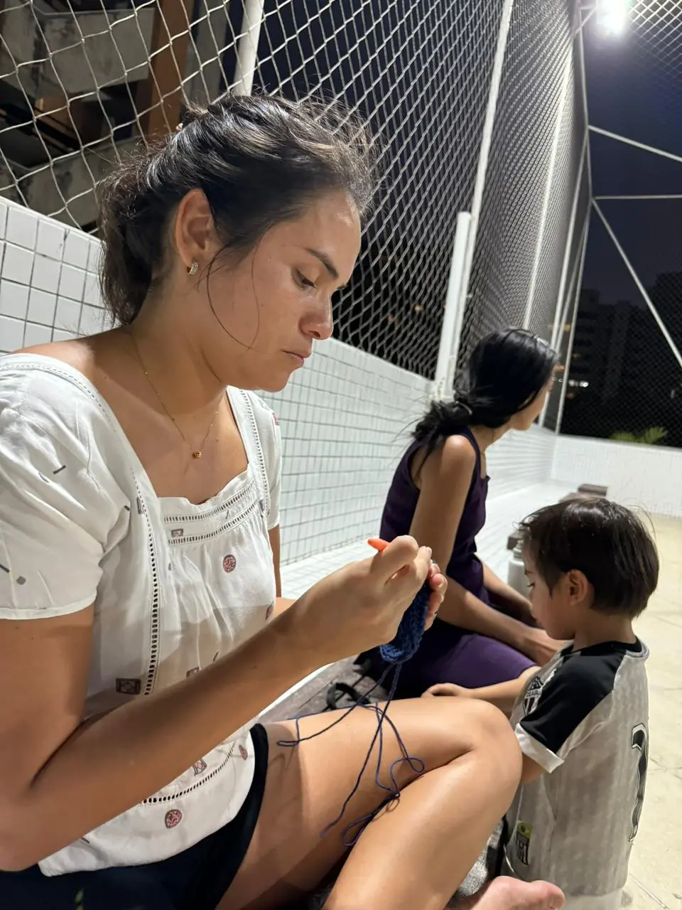 Na imagem, mulher sentada, Cinara Eufrásio, com cabelos castanhos presos, vestindo uma blusa branca e shorts pretos, concentrada em fazer crochê com um fio azul escuro e um gancho laranja. Ao seu lado direito, um menino de camisa cinza e preta olha para a peça de crochê. Ao fundo, uma segunda mulher de vestido roxo está sentada. Eles estão em uma área externa, possivelmente uma varanda ou quadra, com uma tela de proteção e edifícios ao fundo à noite.