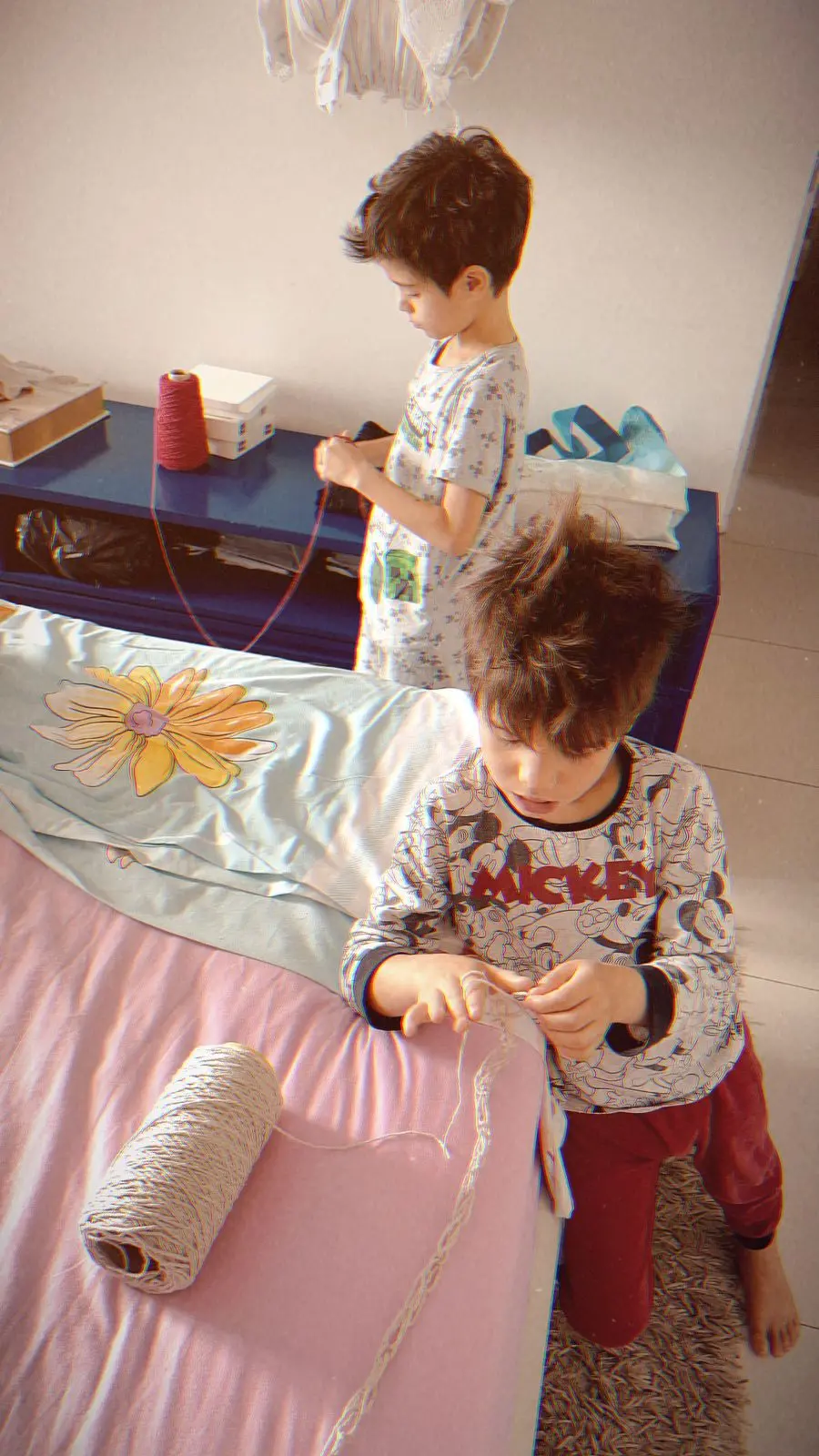 Na imagem, dois meninos estão ajoelhados perto de uma cama e uma cômoda azul, engajados em um projeto de artesanato. O menino em primeiro plano, que veste uma blusa do Mickey, manipula um fio de cor creme que vem de um grande rolo no colchão. O segundo menino, no fundo, também segura um fio vermelho de um carretel vermelho sobre a cômoda.