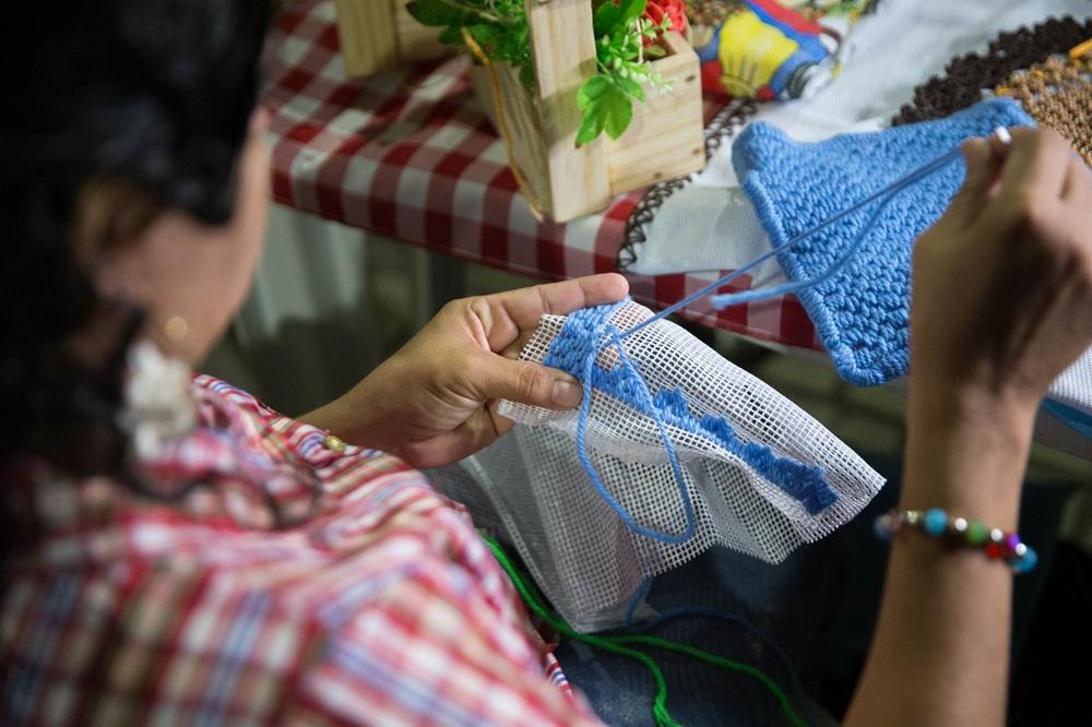 Na imagem, close-up sobre o ombro de uma mulher com cabelos escuros presos, vestindo uma camisa xadrez em tons de vermelho e branco. Ela está usando um gancho de crochê para bordar ou fazer crochê com fio azul brilhante em uma tela de plástico branca com malha aberta. Há uma peça de crochê azul já feita e pequenos objetos de decoração de madeira e flores falsas sobre uma toalha xadrez vermelha e branca ao fundo.