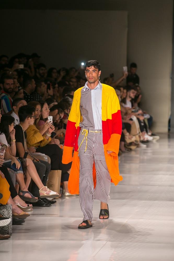 Na imagem, modelo masculino com cabelo escuro e camisa listrada (branca, azul e cinza) desfila em uma passarela, vestindo um conjunto de calças listradas verticais e um sobretudo longo de tricô ou crochê em blocos de cores vibrantes: amarelo, vermelho e laranja. A plateia está sentada em duas fileiras escuras, observando e tirando fotos.