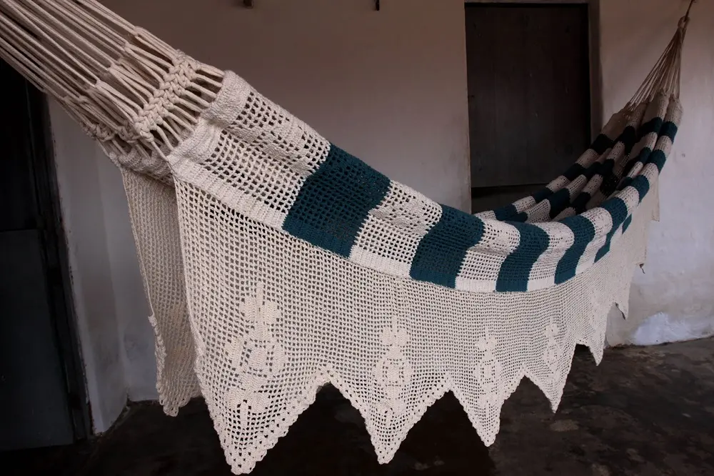 Na imagem, uma rede de dormir (hamaca) artesanal está pendurada em um ambiente interno rústico. A rede é feita predominantemente de crochê em cor creme ou bege claro, com uma faixa horizontal mais larga em blocos de cor verde-petróleo. A borda inferior da rede possui um acabamento de crochê com pontas em formato de bicos, e os punhos são feitos com macramê ou trançado. O fundo é de uma parede clara e um piso escuro.
