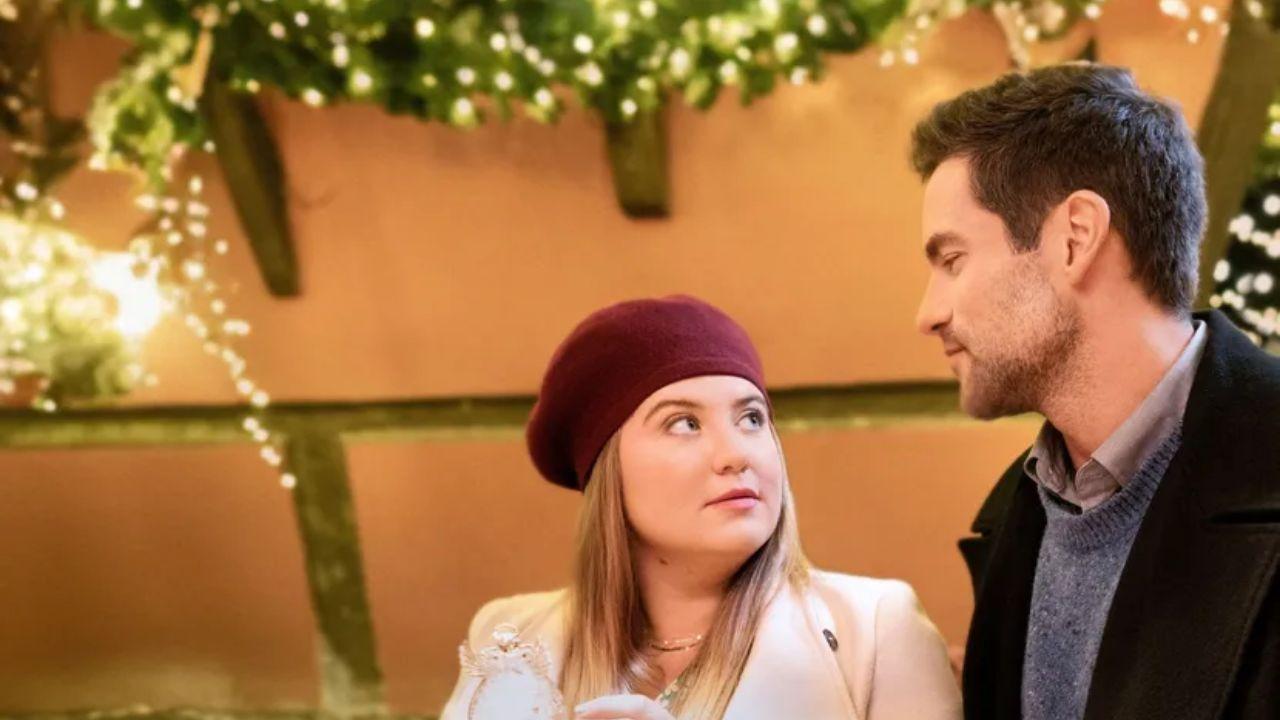 Mulher olhando nos olhos de um homem. Ela está com uma boina na cor bordô e ele estava com um casaco preto. Atrás dos dois, há uma parece com luzes de Natal.