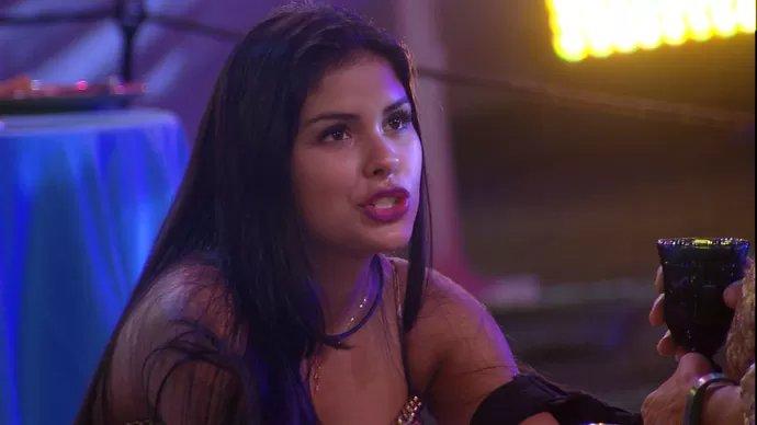 Pessoa com cabelo longo e escuro, usando roupa com detalhes brilhantes e ombros à mostra, segurando um copo escuro em uma cena iluminada por luzes coloridas. O ambiente sugere uma festa ou evento noturno, com mesa coberta por toalha azul ao fundo e iluminação intensa em tons de amarelo e roxo, criando um clima vibrante e festivo.