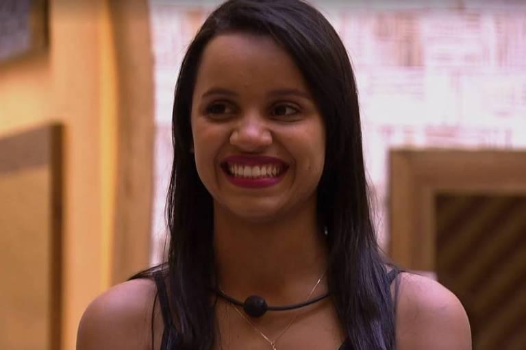 Pessoa com cabelo longo e liso, usando colar preto com detalhe circular e outro colar delicado com pingente pequeno. A roupa é escura e deixa os ombros à mostra. O fundo apresenta uma parede com padrão geométrico em tons claros e uma moldura de madeira, sugerindo um ambiente interno com decoração moderna e acolhedora.