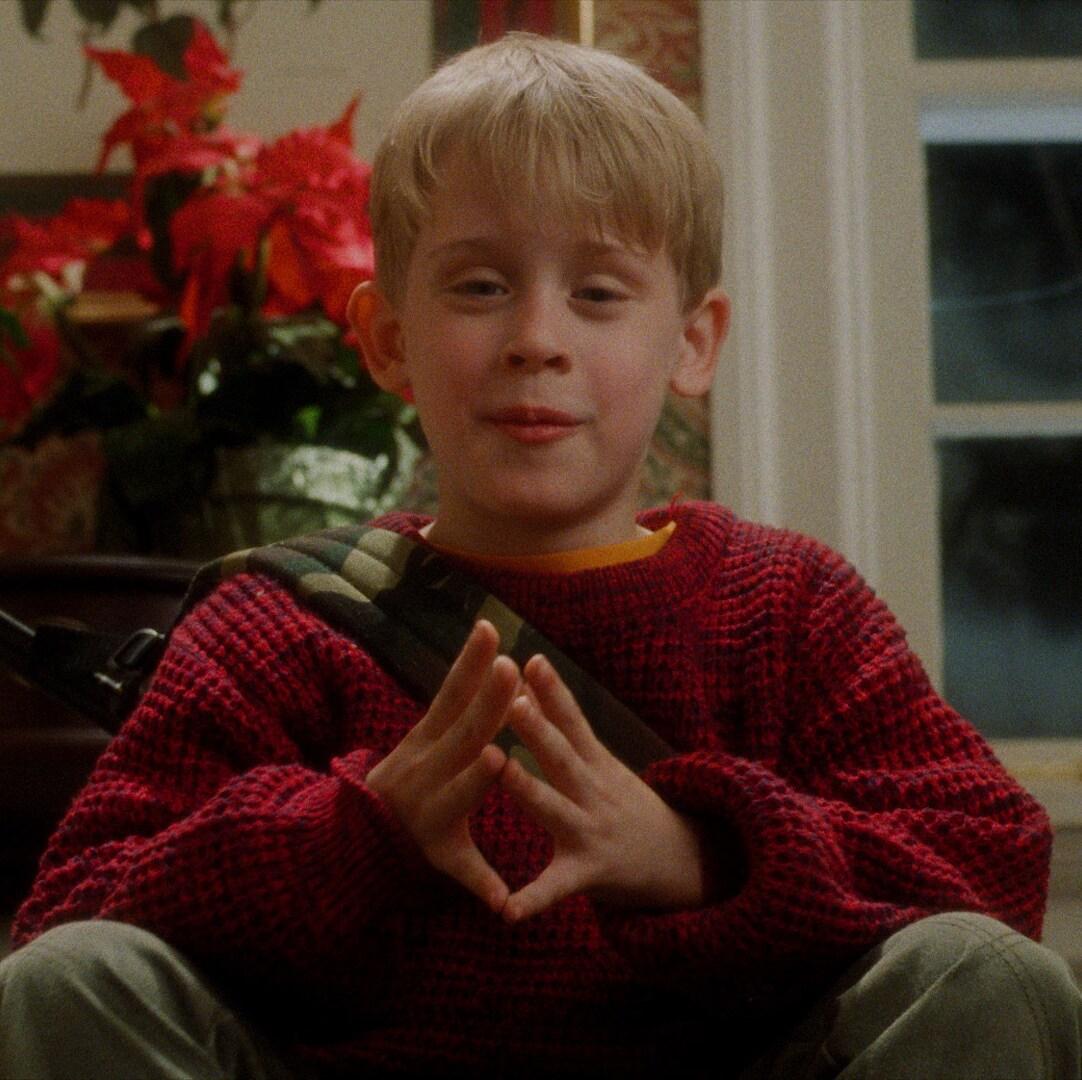 Criança loira vestida com suéter vermelho e levantando as mãos ao peito como se estivesse falando algo.