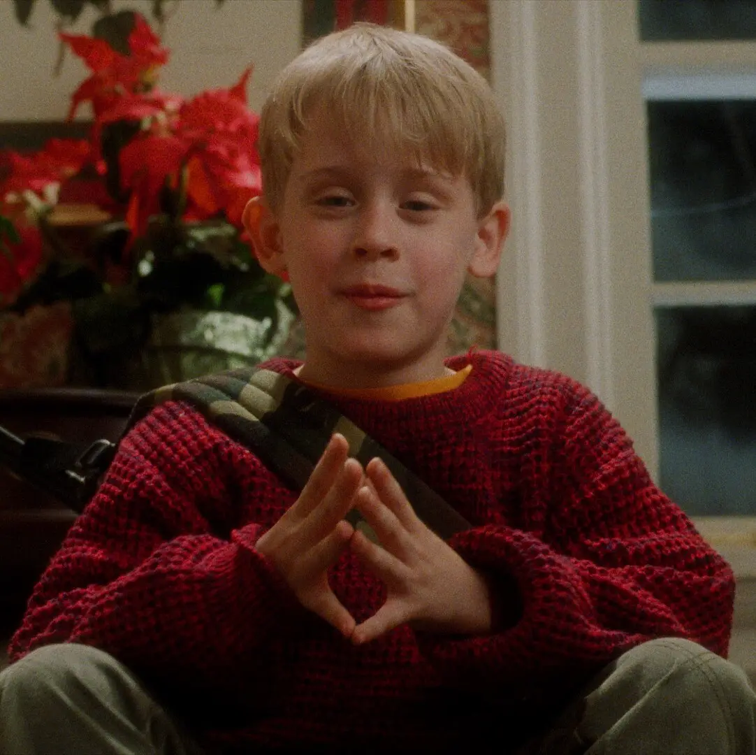 Criança loira vestida com suéter vermelho e levantando as mãos ao peito como se estivesse falando algo.