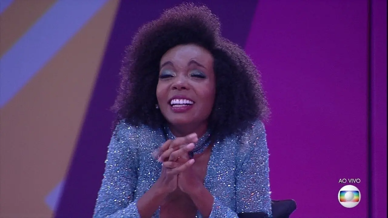 Pessoa com cabelo volumoso e cacheado, vestindo uma roupa brilhante em tom azul claro com textura cintilante. A peça tem decote profundo e mangas longas, transmitindo elegância e sofisticação. O fundo apresenta uma parede roxa com elementos geométricos e iluminação intensa, sugerindo um cenário de programa televisivo ao vivo, indicado pelo texto “AO VIVO” e o logotipo da Globo no canto inferior direito.