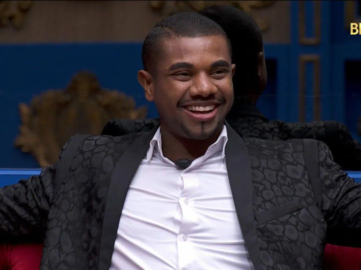 Pessoa vestindo um blazer preto com textura em padrão geométrico discreto e lapelas lisas, combinado com camisa branca por baixo. O look transmite sofisticação e estilo formal, ideal para eventos importantes ou aparições televisivas. O fundo apresenta elementos decorativos clássicos em tons dourados e azuis, sugerindo um cenário elegante e bem iluminado.