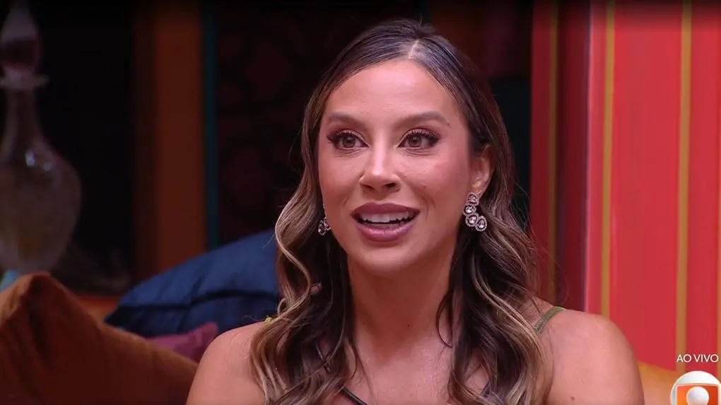 Pessoa com cabelo longo e ondulado, usando brincos grandes e brilhantes com design sofisticado. Veste uma roupa elegante em tom neutro, com alças finas, transmitindo estilo refinado. O cenário apresenta uma decoração luxuosa com parede em tom vermelho vibrante e detalhes dourados, além de almofadas escuras ao fundo e iluminação suave, sugerindo um ambiente interno acolhedor e sofisticado.