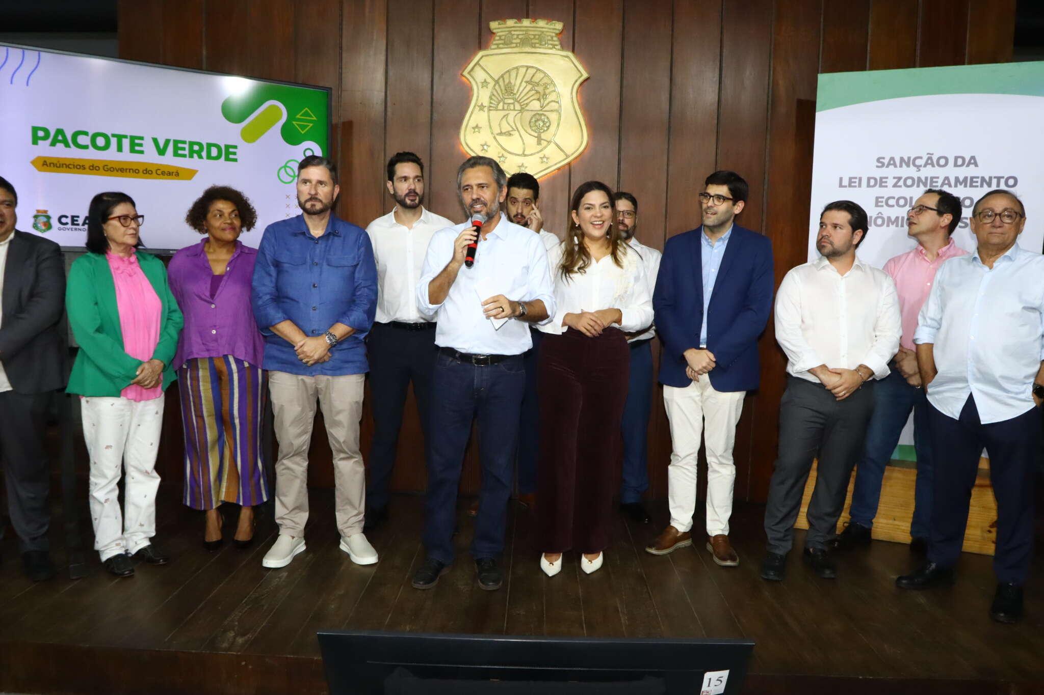 Um grupo de cerca de treze pessoas está em pé sobre um palco, diante de um painel de madeira com o brasão do Estado do Ceará ao centro. No meio do grupo, um homem segura um microfone e fala, enquanto os demais posam para a foto. À esquerda, há uma tela com o texto “Pacote Verde – Anúncios do Governo do Ceará”. À direita, outro painel exibe o texto “Sanção da Lei de Zoneamento Ecológico-Econômico”. As pessoas usam roupas formais ou semiformal, e parecem participar de um evento oficial do governo.