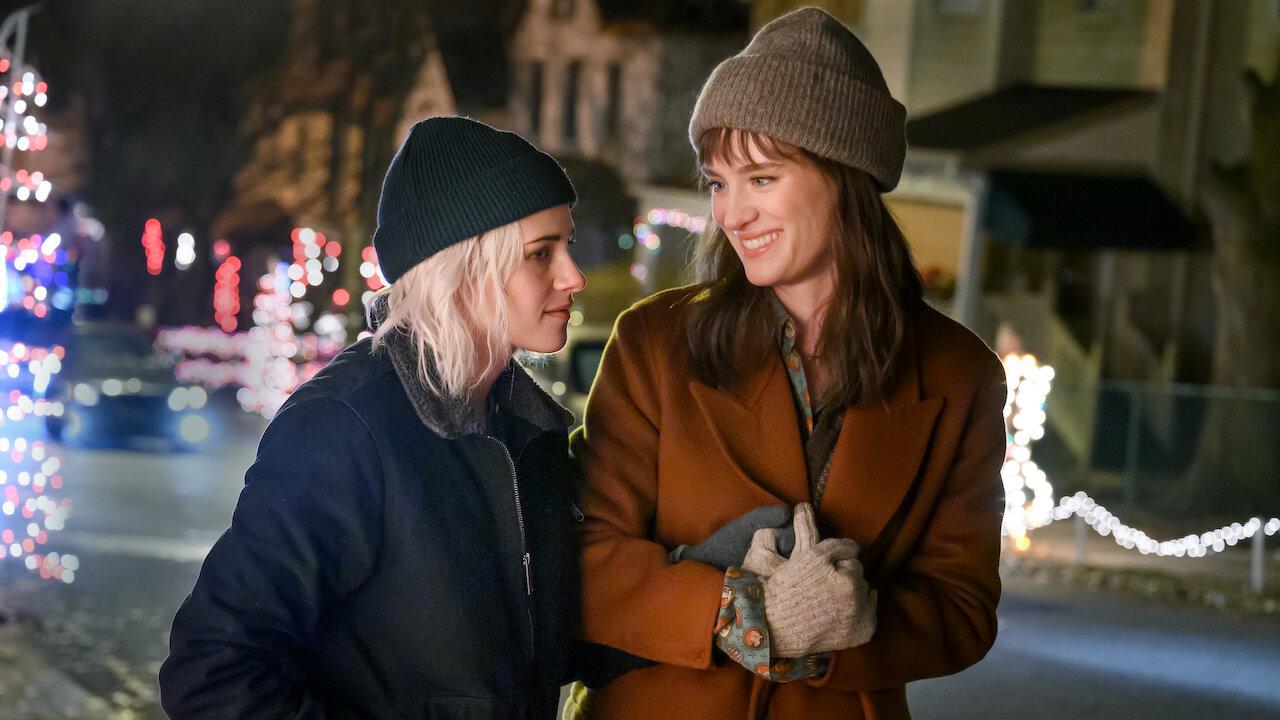 Cena noturna com decoração natalina: duas pessoas com roupas de inverno em uma rua iluminada por luzes festivas.