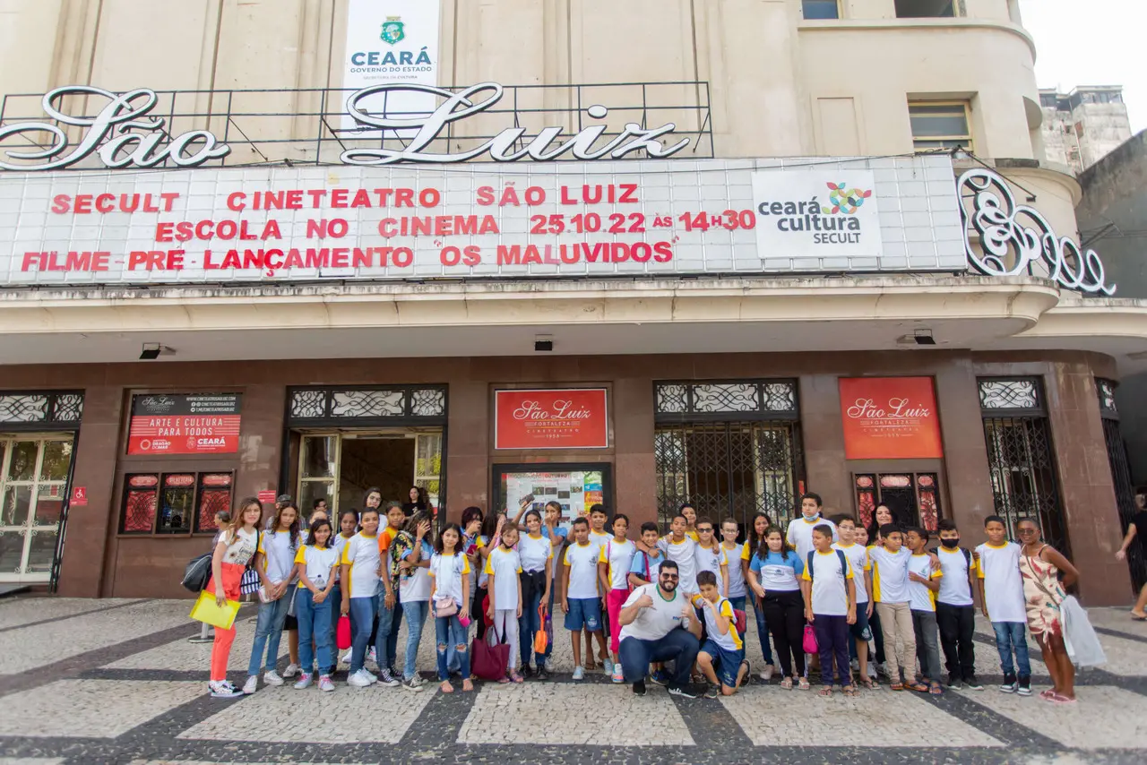 Uma foto tirada de frente mostra um grupo de estudantes e adultos posando na frente da fachada do Cineteatro São Luiz, em Fortaleza.