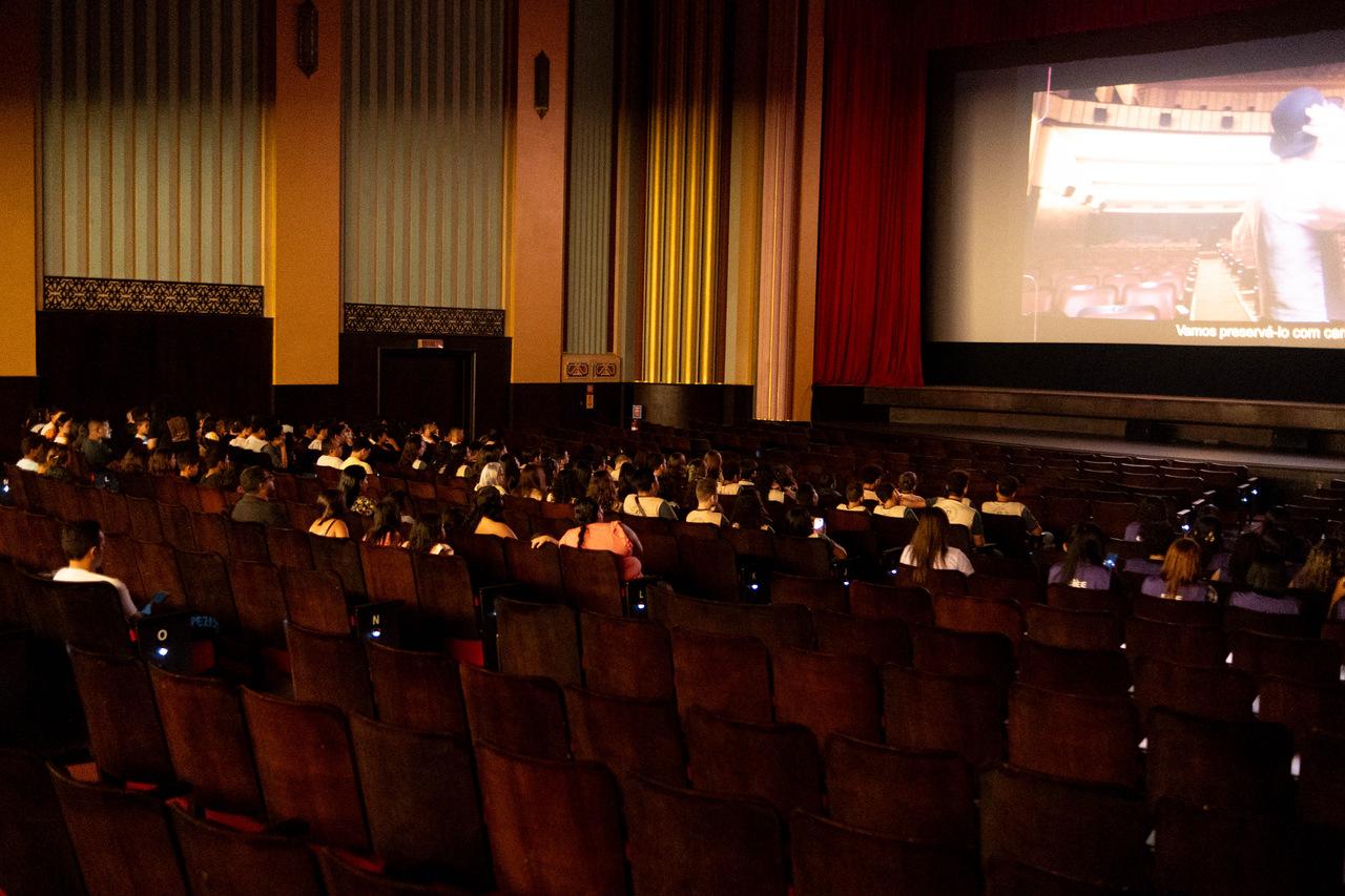Uma foto interna, em plano aberto e em baixa luminosidade, capta o público assistindo a uma exibição no salão principal do Cineteatro São Luiz.