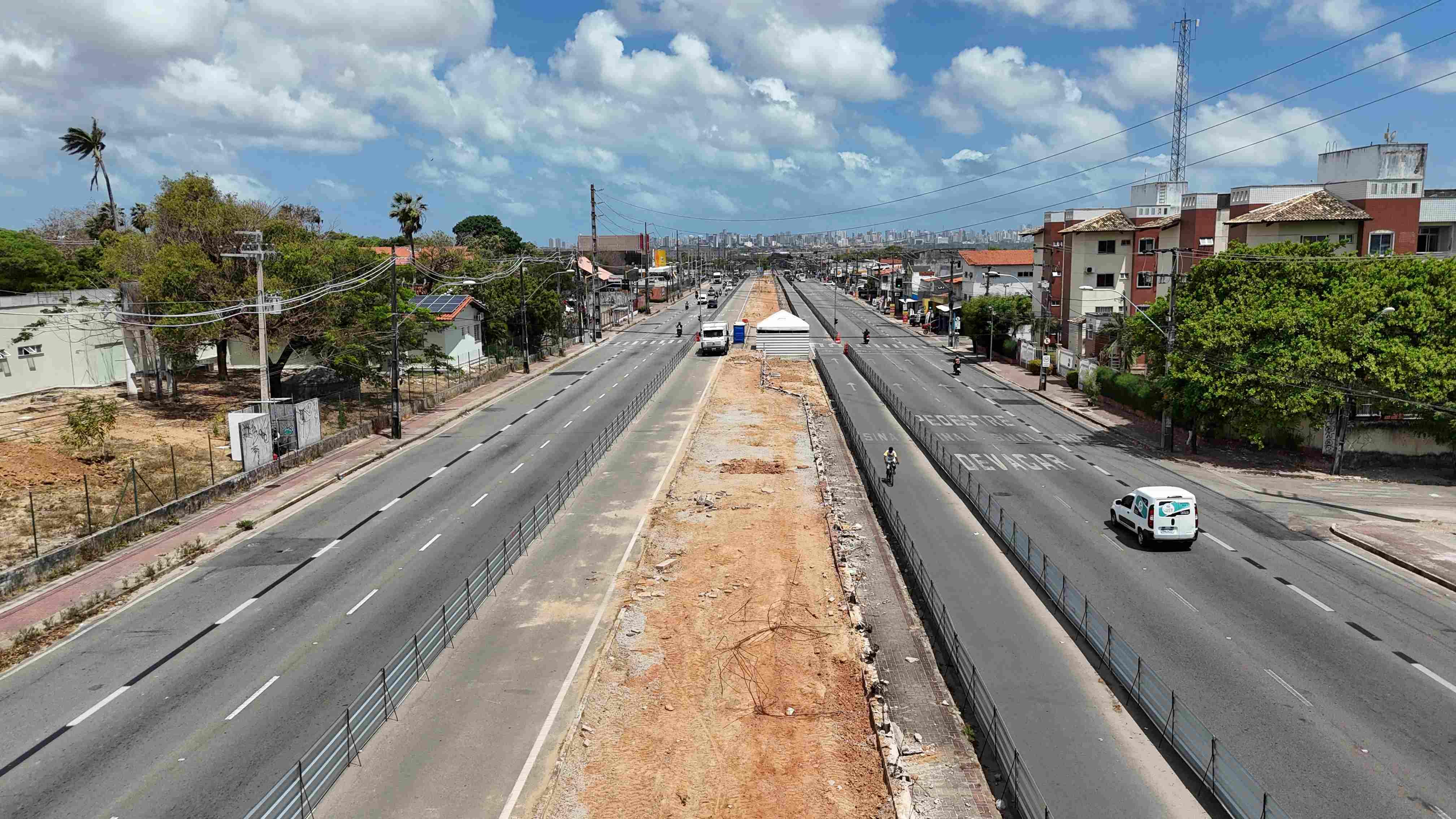 Imagem de uma via urbanizada em construção, com múltiplas faixas de trânsito, árvores ao lado e um céu parcialmente nublado, em uma área de cidade.