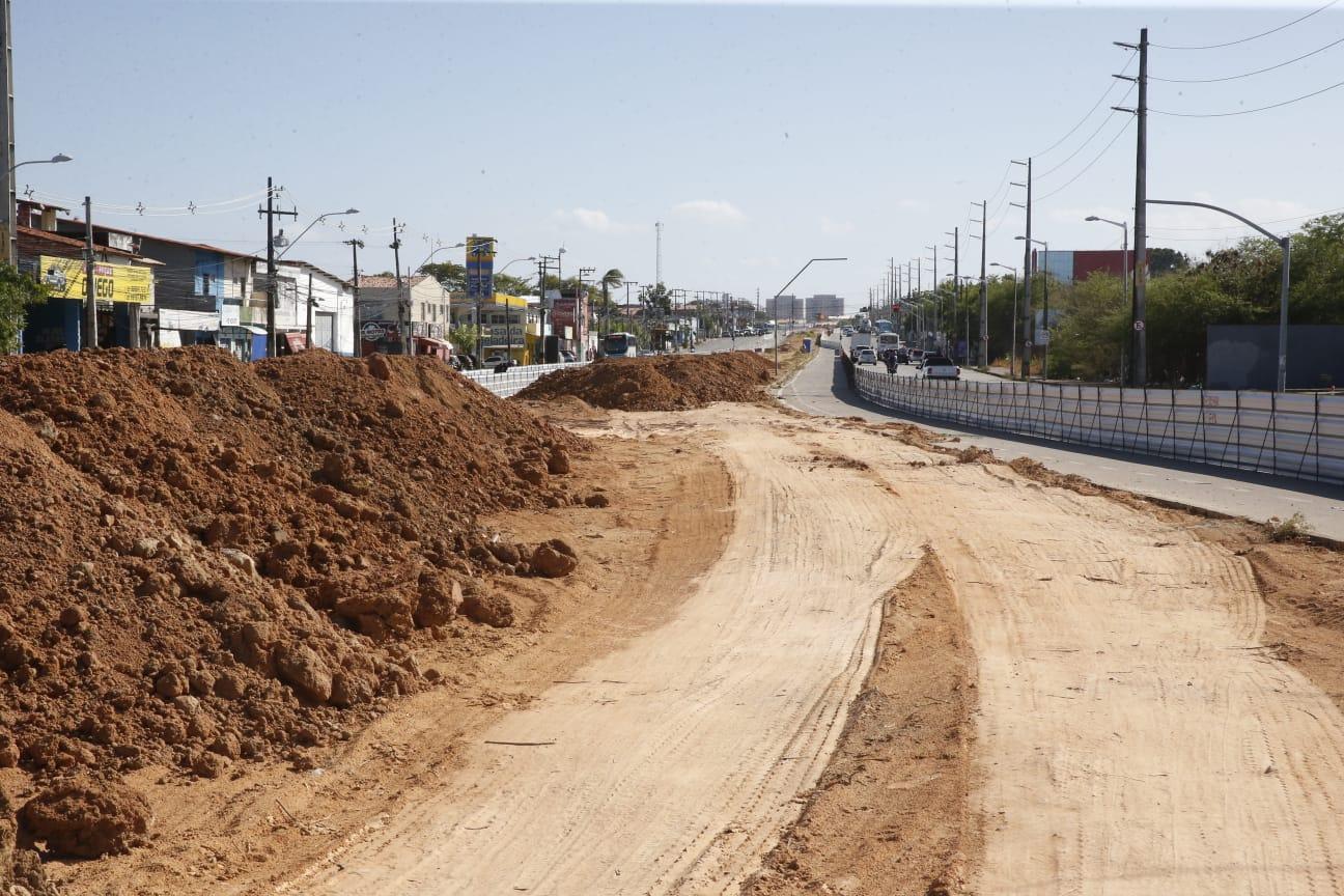 Obras de construção e melhorias na infraestrutura de uma estrada urbana, com terra e brita sendo movimentadas, possível recuperação de vias urbanas.
