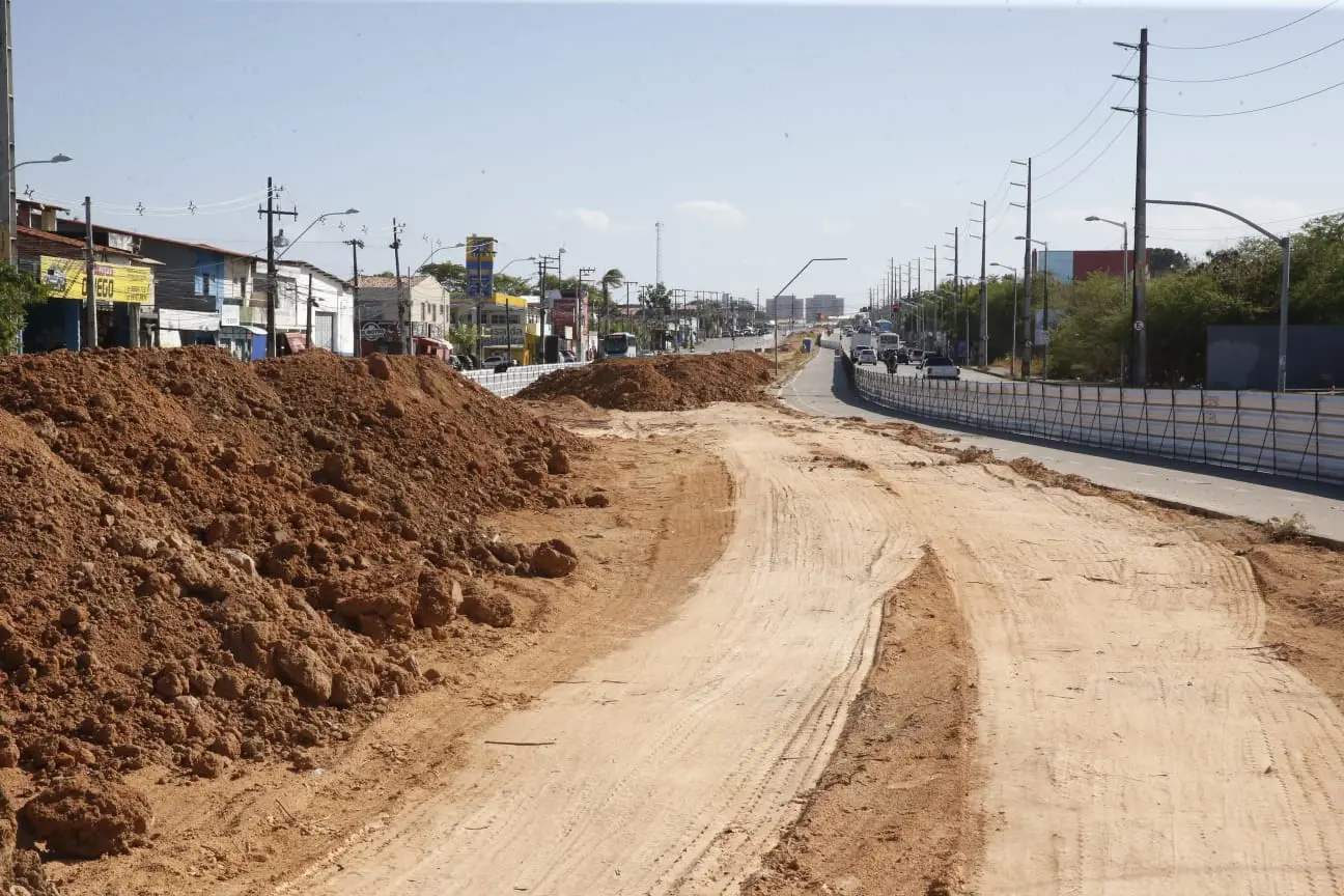 Obras de construção e melhorias na infraestrutura de uma estrada urbana, com terra e brita sendo movimentadas, possível recuperação de vias urbanas.