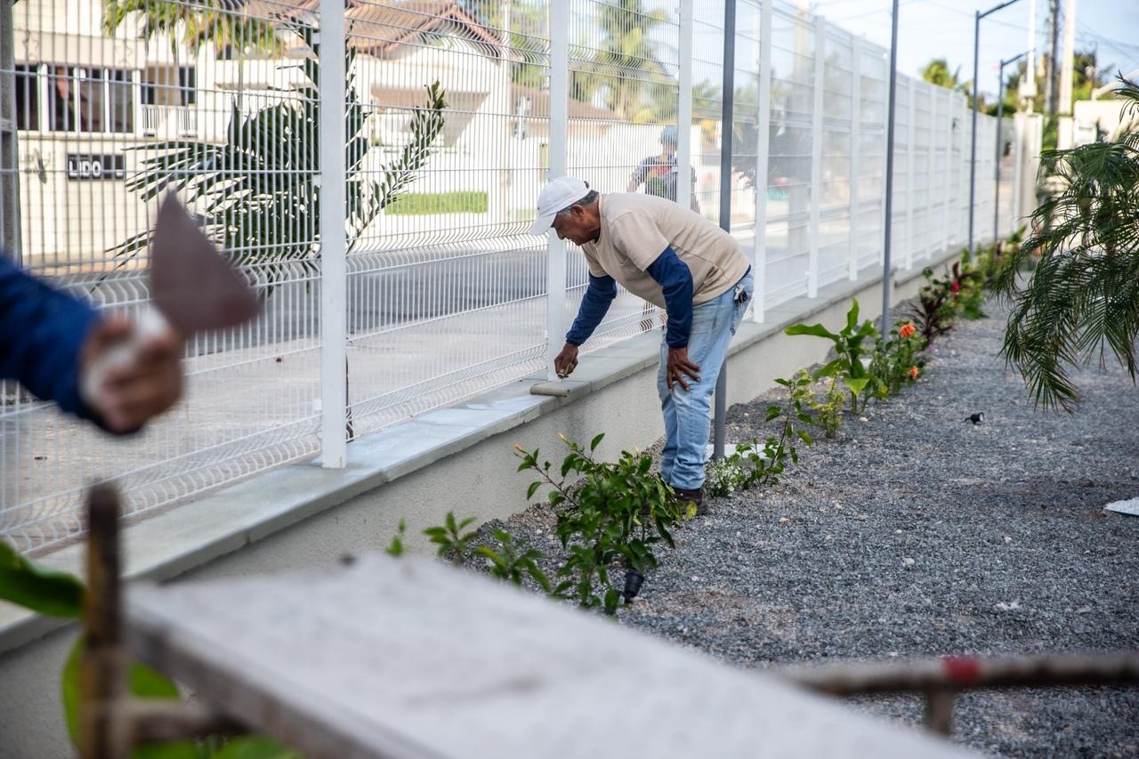 Um trabalhador, vestindo camisa bege e boné branco, está abaixado pintando de branco o rodapé de um muro de concreto sob uma cerca de metal branca. O chão é coberto por brita, com pequenas mudas e plantas ao longo do muro.