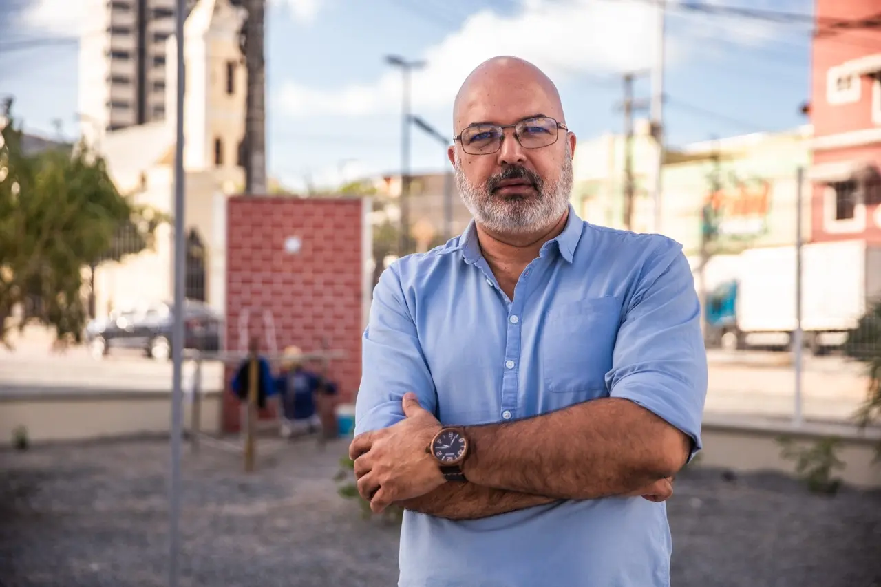 Retrato de Sandro Gouveia, pró-reitor de Cultura da UFC, um homem careca, com barba grisalha e óculos, vestindo camisa social azul clara, posando de braços cruzados. O fundo urbano está desfocado, mostrando um muro de tijolos vermelhos e edifícios.