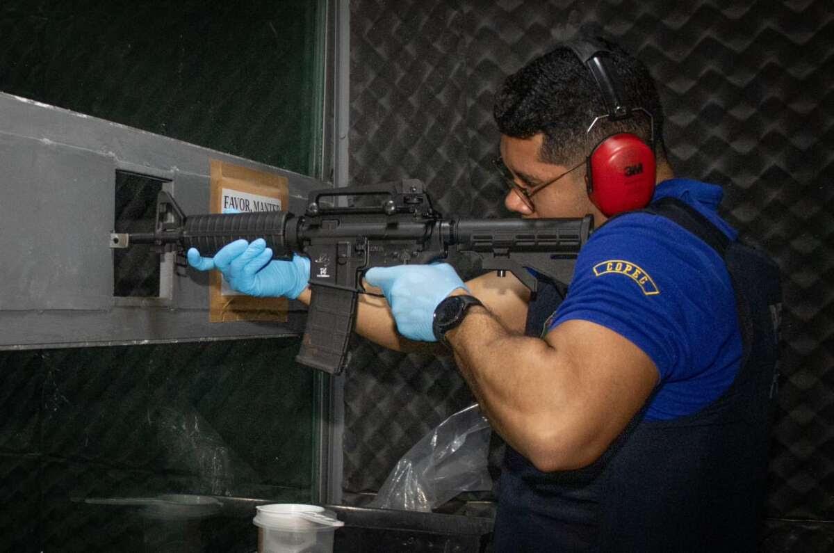 A imagem mostra um perito forense, com farda azul, luvas e fone de ouvido, realizando um teste com uma arma de fogo de grande porte.