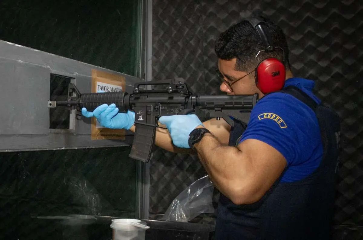 A imagem mostra um perito forense, com farda azul, luvas e fone de ouvido, realizando um teste com uma arma de fogo de grande porte.