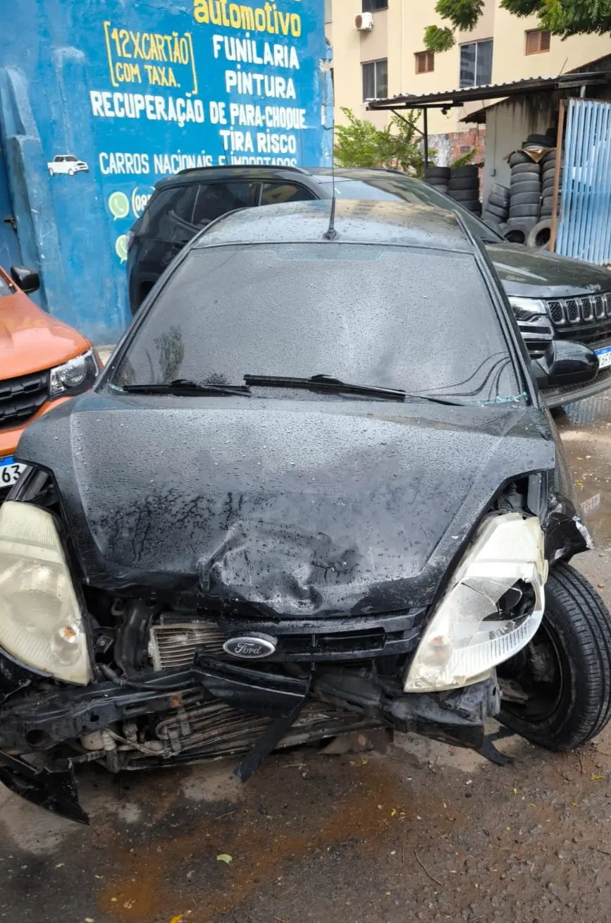 Carro preto com a frente destruída após acidente, com capô amassado, faróis quebrados e para-choque danificado, estacionado em frente a uma oficina de funilaria e pintura.