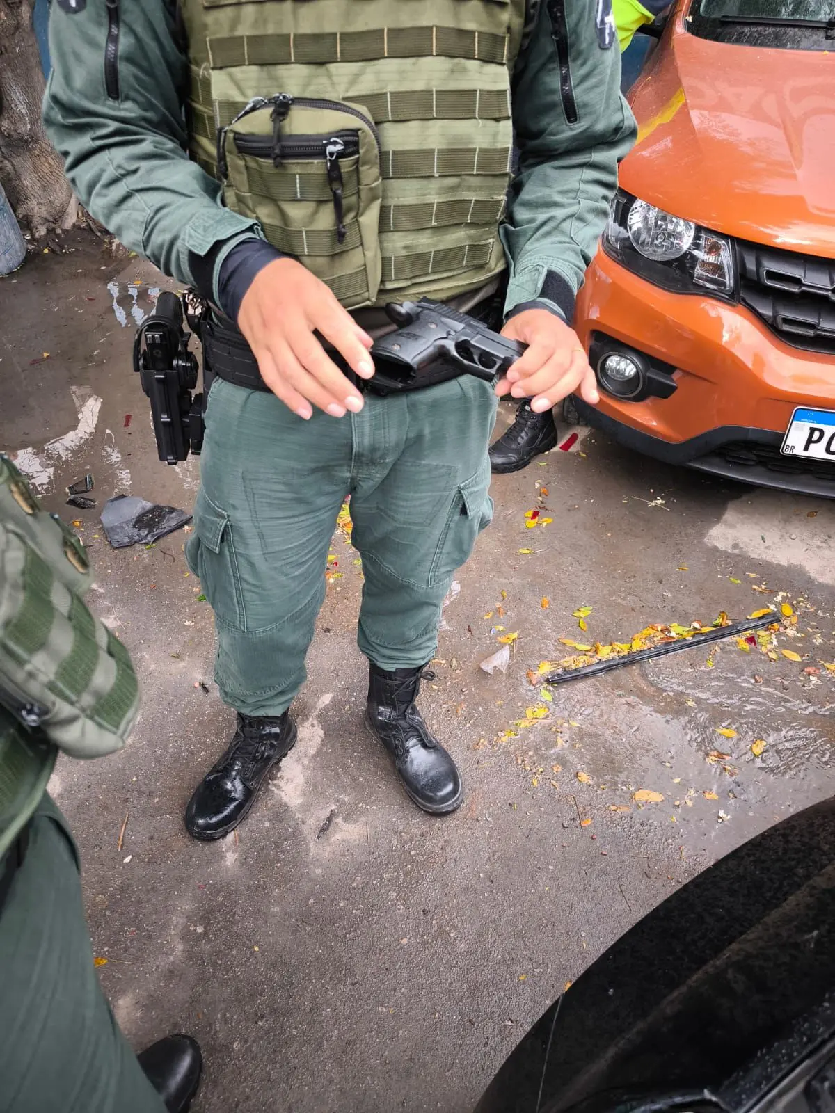 Agente de segurança com uniforme verde segura um simulacro de arma de fogo na altura da cintura, em via pública, com viatura estacionada ao fundo.