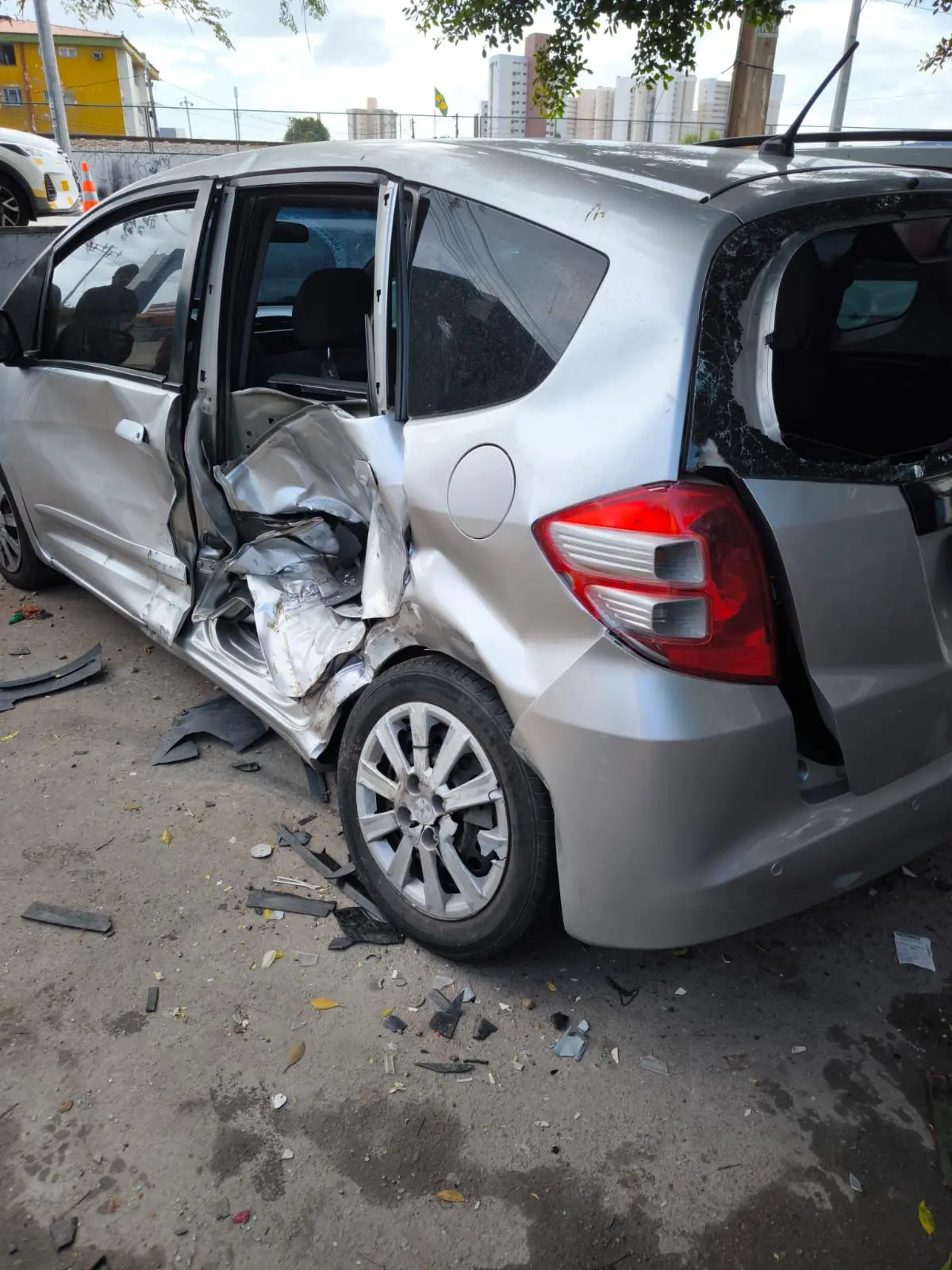 Carro hatch prateado com forte avaria na lateral esquerda e vidros quebrados após colisão, estacionado em via urbana com destroços no chão.