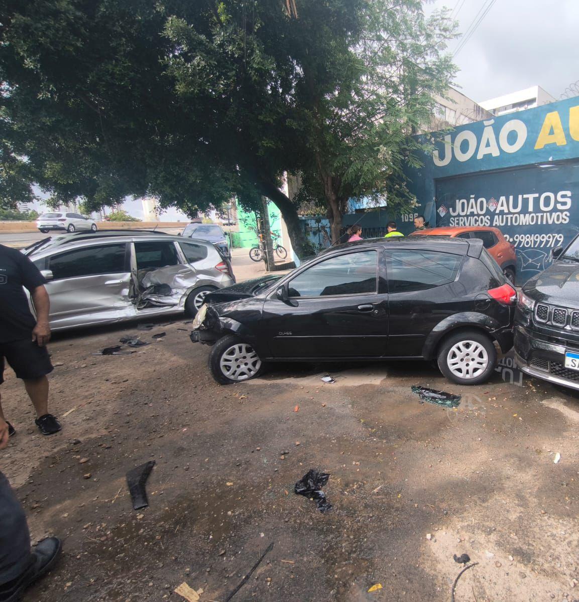 Carros danificados após colisão em via urbana; veículo preto com a frente amassada e carro prateado com a lateral destruída, enquanto pessoas observam o acidente próximo a uma oficina automotiva.