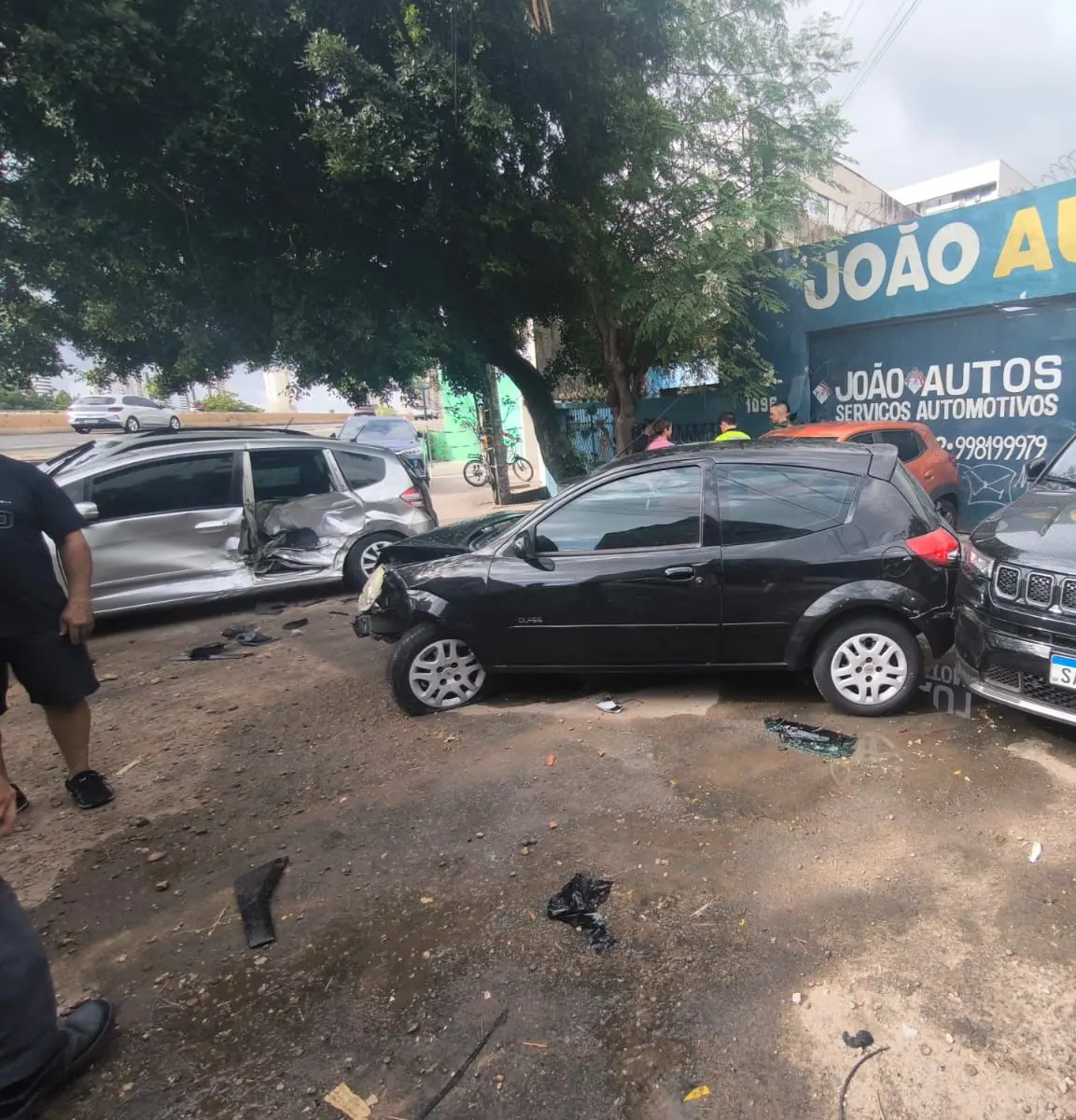 Carros danificados após colisão em via urbana; veículo preto com a frente amassada e carro prateado com a lateral destruída, enquanto pessoas observam o acidente próximo a uma oficina automotiva.