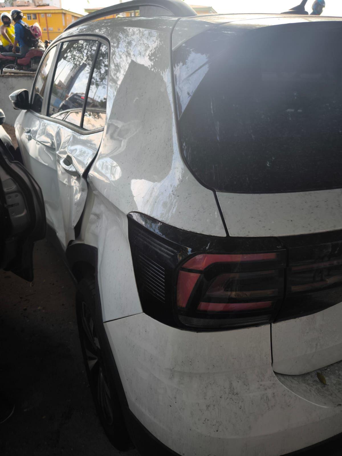 Carro branco com amassado na lateral traseira esquerda após colisão, estacionado em via urbana, com outros veículos e motociclistas ao fundo.