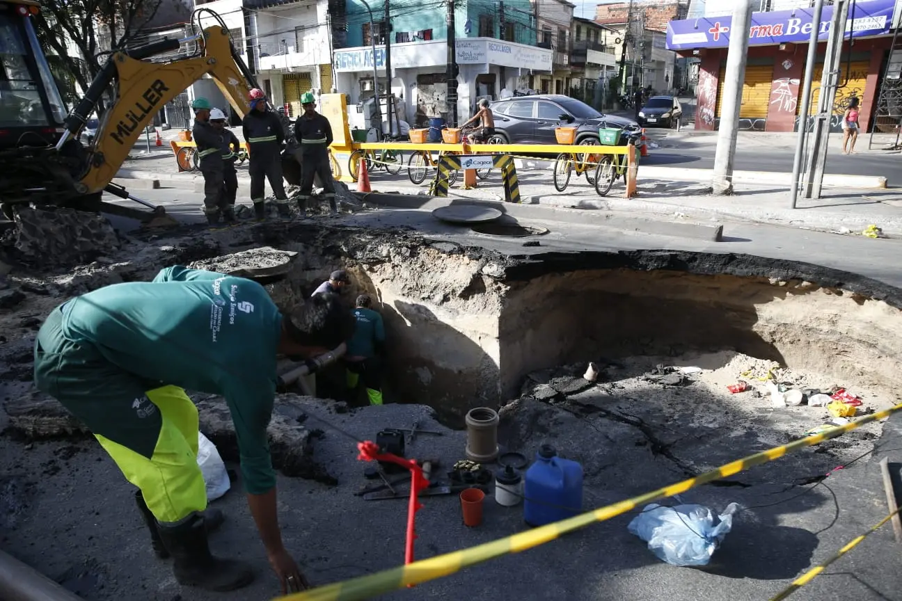 Imagem mostra avenida com cratera formada em buraco, ocupando todo trecho da via. Trabalhadores fazem obras de reparo no local.