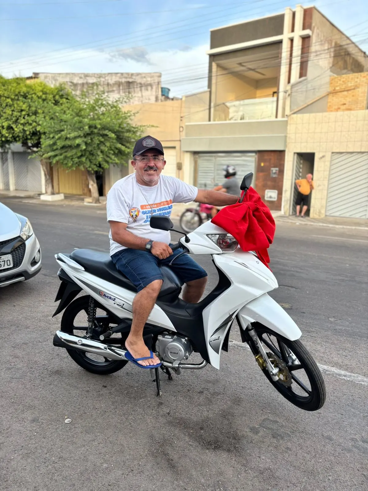 Homem sorridente com camiseta branca, chapéu e chinelos, sentado em uma motocicleta branca na rua, com prédio residencial ao fundo e uma pessoa ao lado.