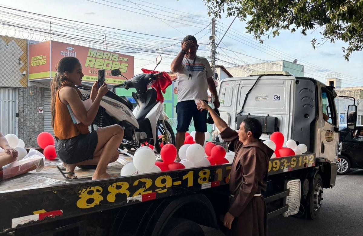 Homem celebrando presente de moto, balões vermelhos e brancos, em uma rua da cidade, com uma pessoa segurando um celular para tirar foto.