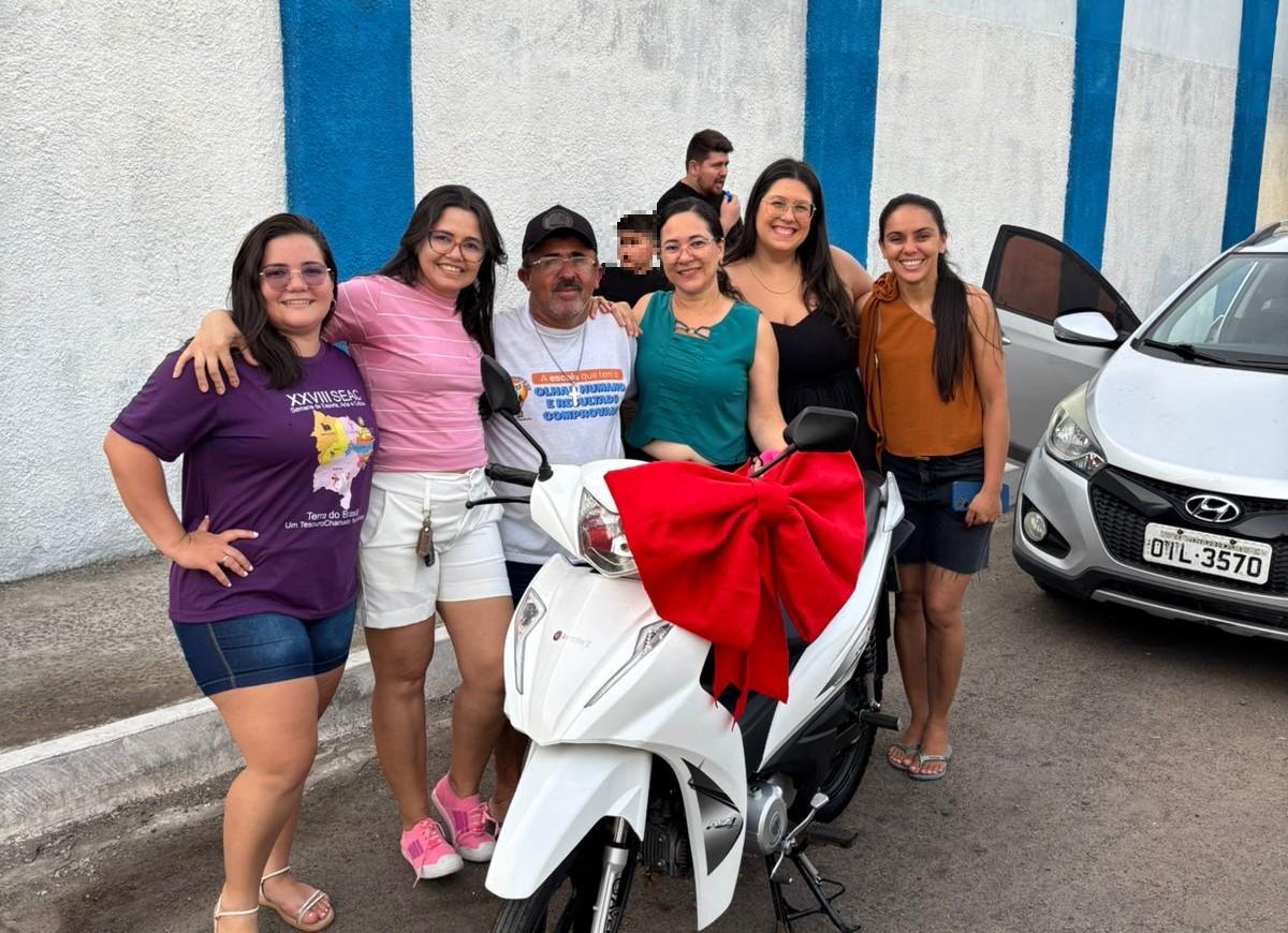 Grupo de amigos comemorando com uma motocicleta decorada com laço vermelho, ao lado de carros na rua.