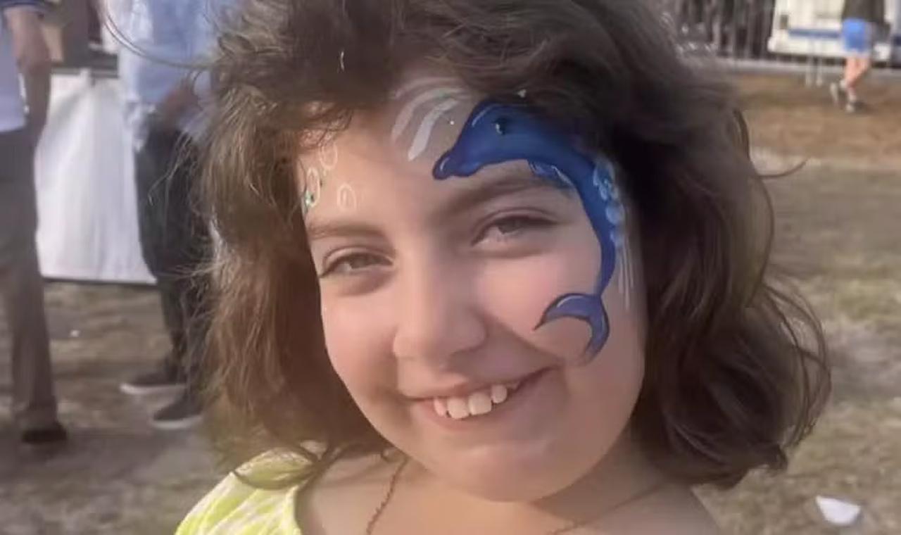 Criança branca com pintura de rosto de um golfinho azul. Ela tem cabelos castanhos curtos e sorri para a foto.