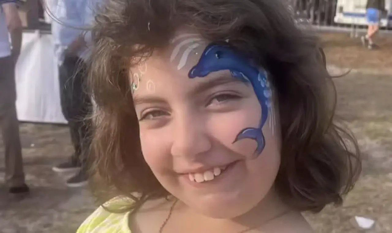 Criança branca com pintura de rosto de um golfinho azul. Ela tem cabelos castanhos curtos e sorri para a foto.