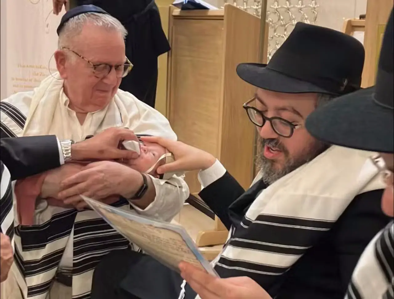 Rabino de barba castanha e chapeu coco preto sentado lendo para um senhor de cabelo branco e blusa branca.