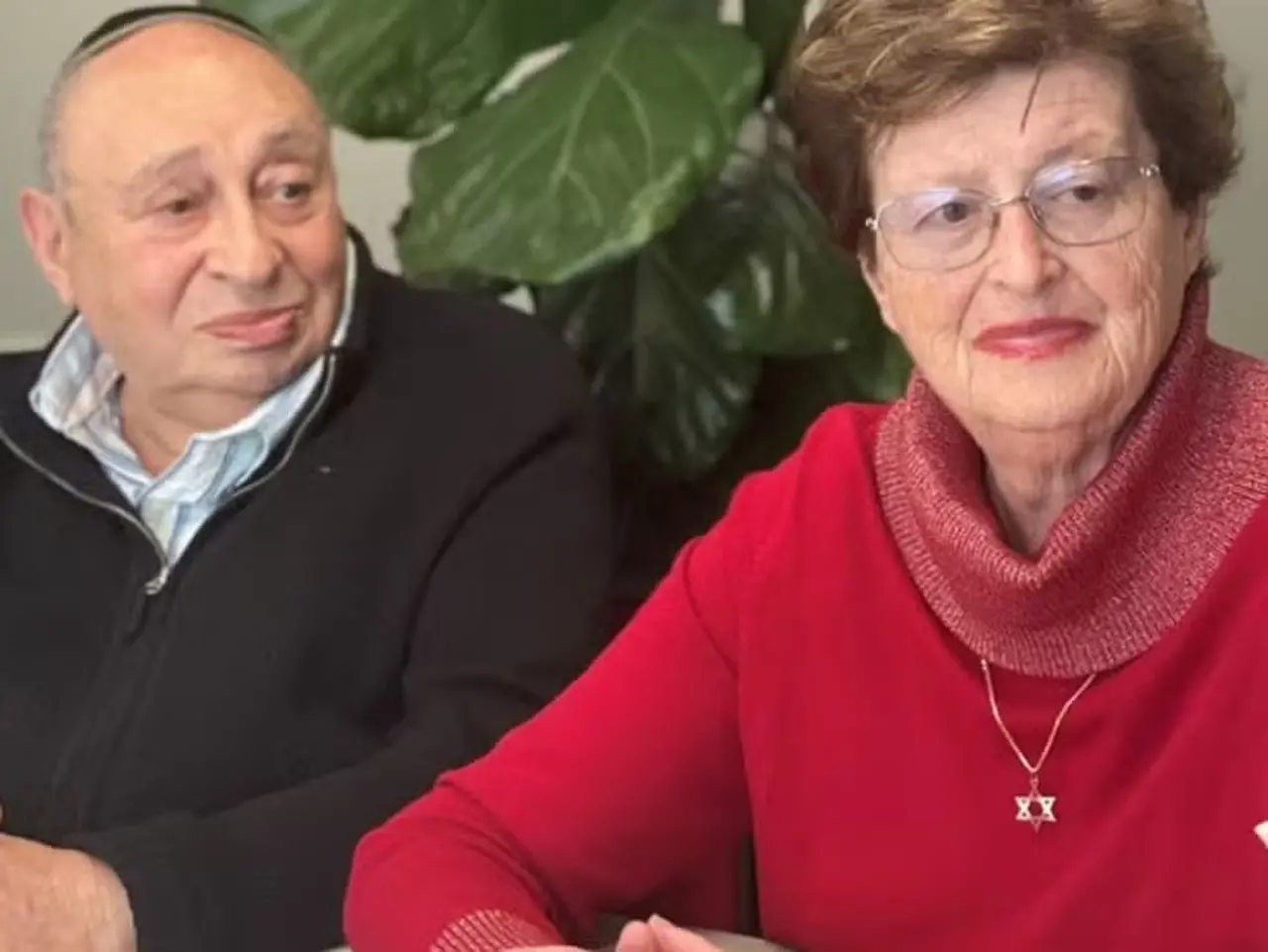 Casal de idosos brancos. O homem é quase careca e veste blusa longa preta, e a mulher tem cabelo curto castanho, usa óculos e veste roupa vermelha.