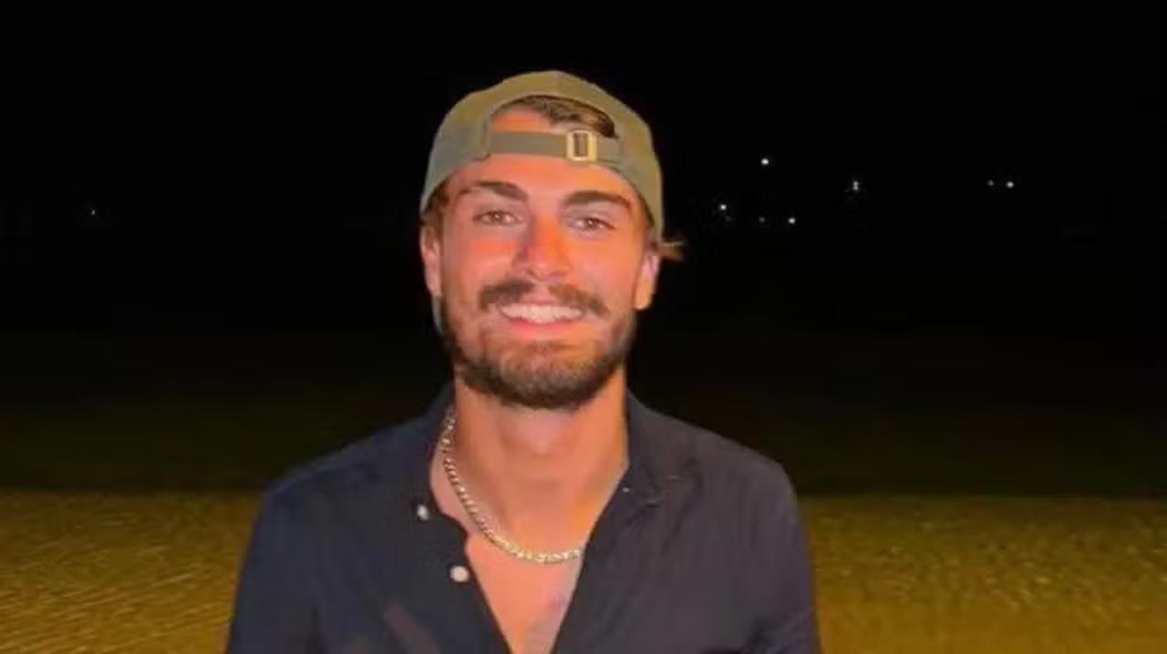 Jovem branco de barba e bigode castanhos sorrindo para foto. Ele veste blusa desabotoada azul marinho e boné verde para trás.