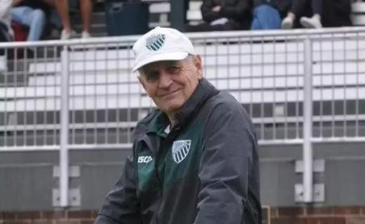 Homem branco de boné branco com escudo de time preto e branco e jaqueta preta com mesmo símbolo.