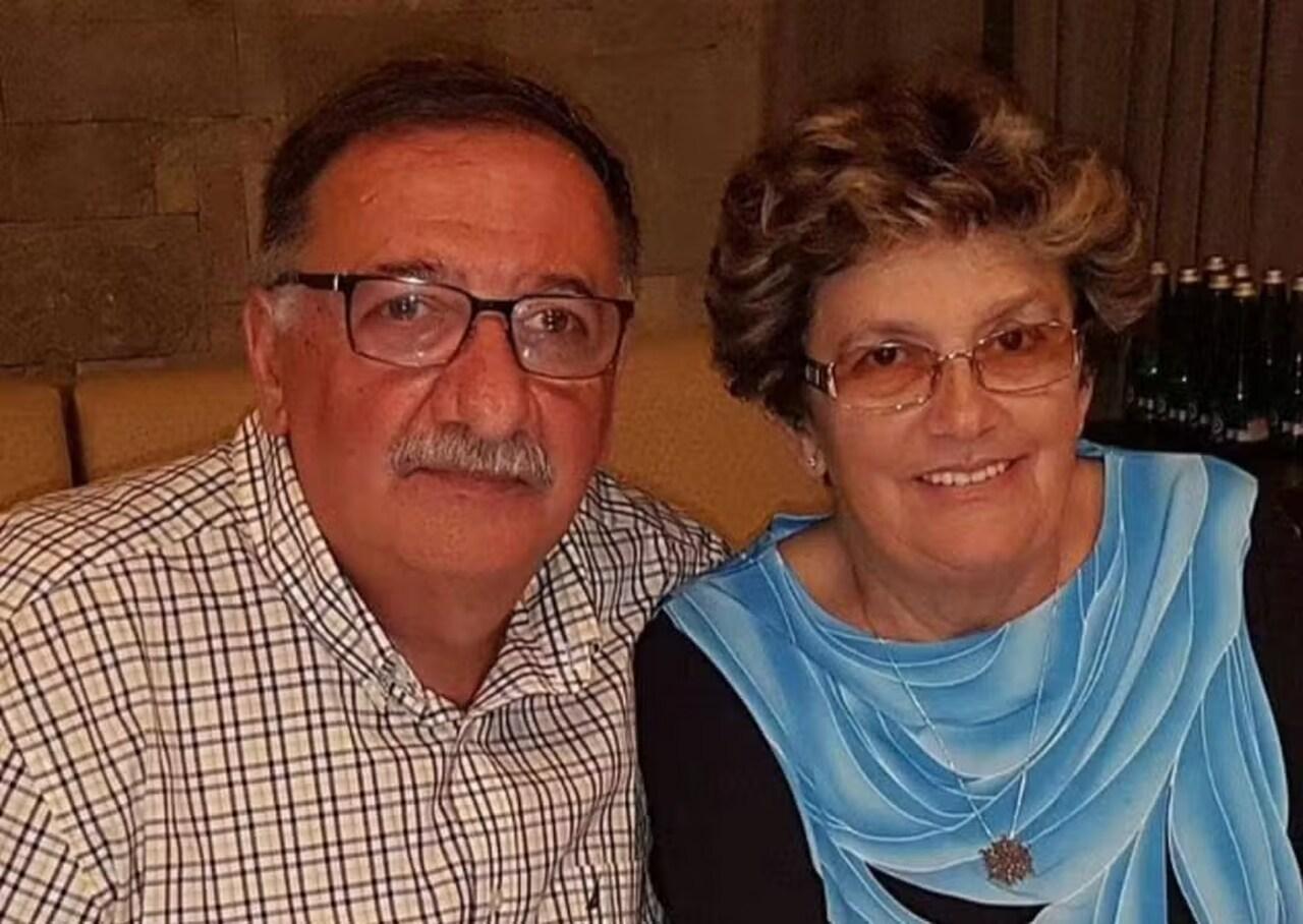 Casal da terceira idade brancos. Homem tem bigode branco e usa óculos e camisa quadriculada preta e branca. Mulher tem cabelos curtos e louros, usa óculos e está com blusa azul de mangas pretas.