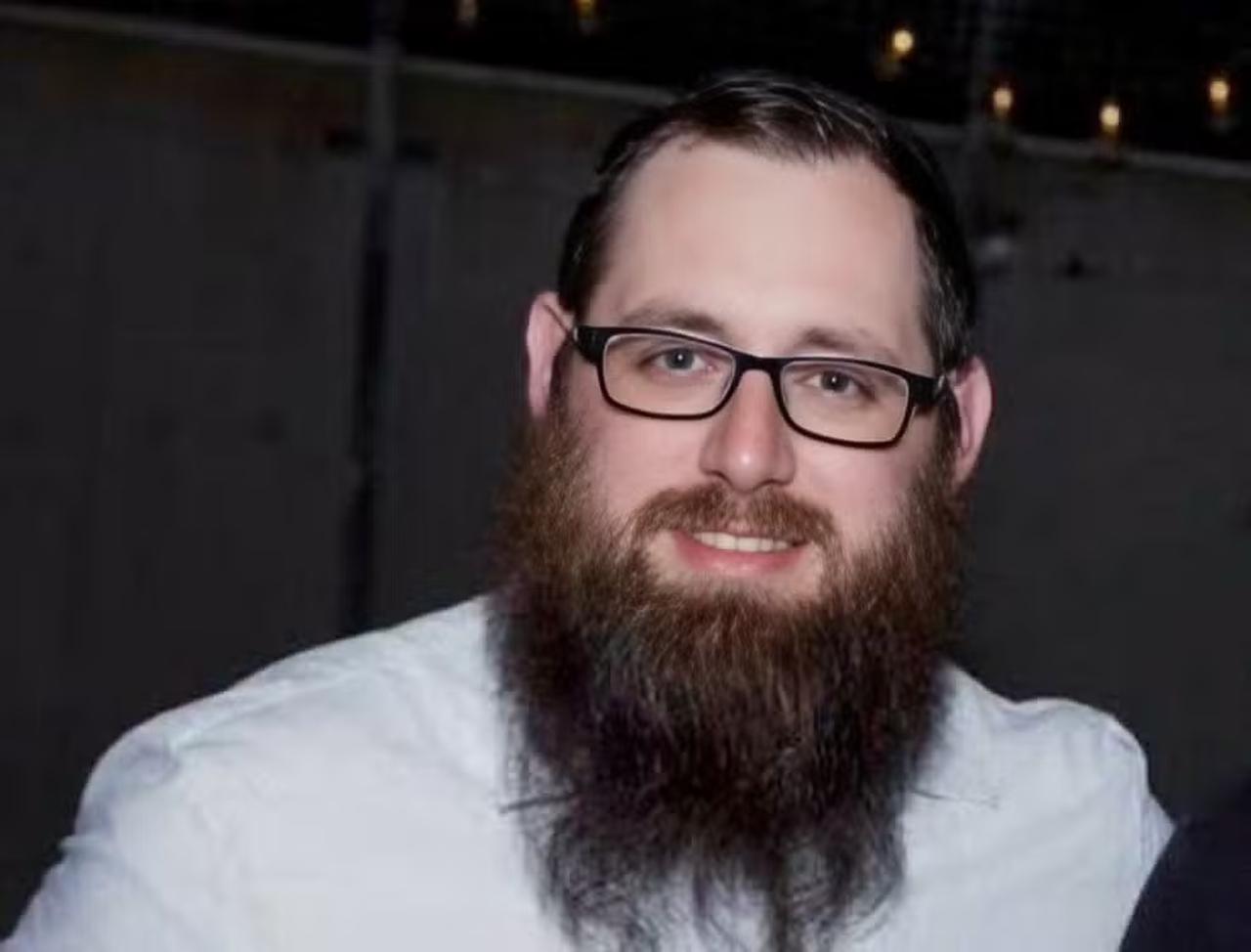 Homem branco de óculos, barba longa e cabelo castanho, usando blusa branca e sorrindo para a foto.