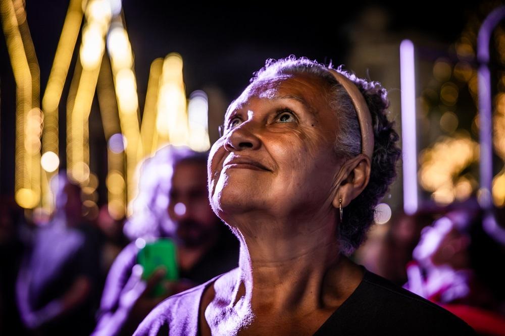 Na imagem, uma foto noturna e de close-up no rosto de uma mulher idosa de pele clara e cabelo grisalho, Dona Marilene. Ela está olhando para cima e ligeiramente para a esquerda com uma expressão de encantamento ou admiração. O rosto dela está iluminado por uma luz quente (laranja/amarela). Ela usa uma faixa clara no cabelo e um brinco pequeno. O fundo está em bokeh (desfocado), mostrando luzes amarelas intensas em formatos de arco (possivelmente decorações de Natal) e sombras de pessoas, algumas com tons arroxeados devido à iluminação ambiente.