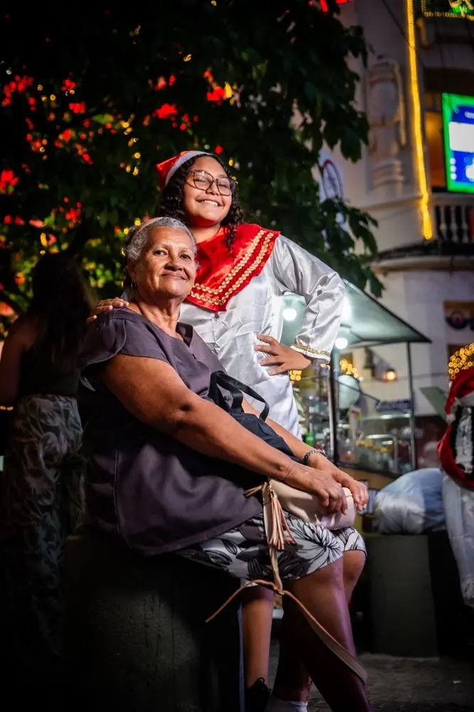 Na imagem, uma foto noturna e vertical em ambiente externo. Em primeiro plano, sentada em um banco ou assento escuro, está Dona Marilene, uma mulher idosa de pele clara e cabelo grisalho, sorrindo para a câmera. Ela usa uma blusa escura de manga curta e uma saia estampada clara. Ela tem uma bolsa preta apoiada na perna. Atrás dela, em pé, está Ana Emilly, uma jovem de pele morena, cabelo cacheado e óculos, com um sorriso alegre e a mão na cintura. Ela veste um traje de Natal composto por uma bata branca de cetim com detalhes dourados e um xale vermelho e branco, além de um gorro de Papai Noel. Elas estão abraçadas, com o braço de Dona Marilene no ombro de Ana Emilly. O fundo mostra uma árvore escura com luzes de Natal vermelhas, um prédio iluminado em estilo clássico e uma barraca de rua, tudo iluminado por luzes amarelas e brancas.